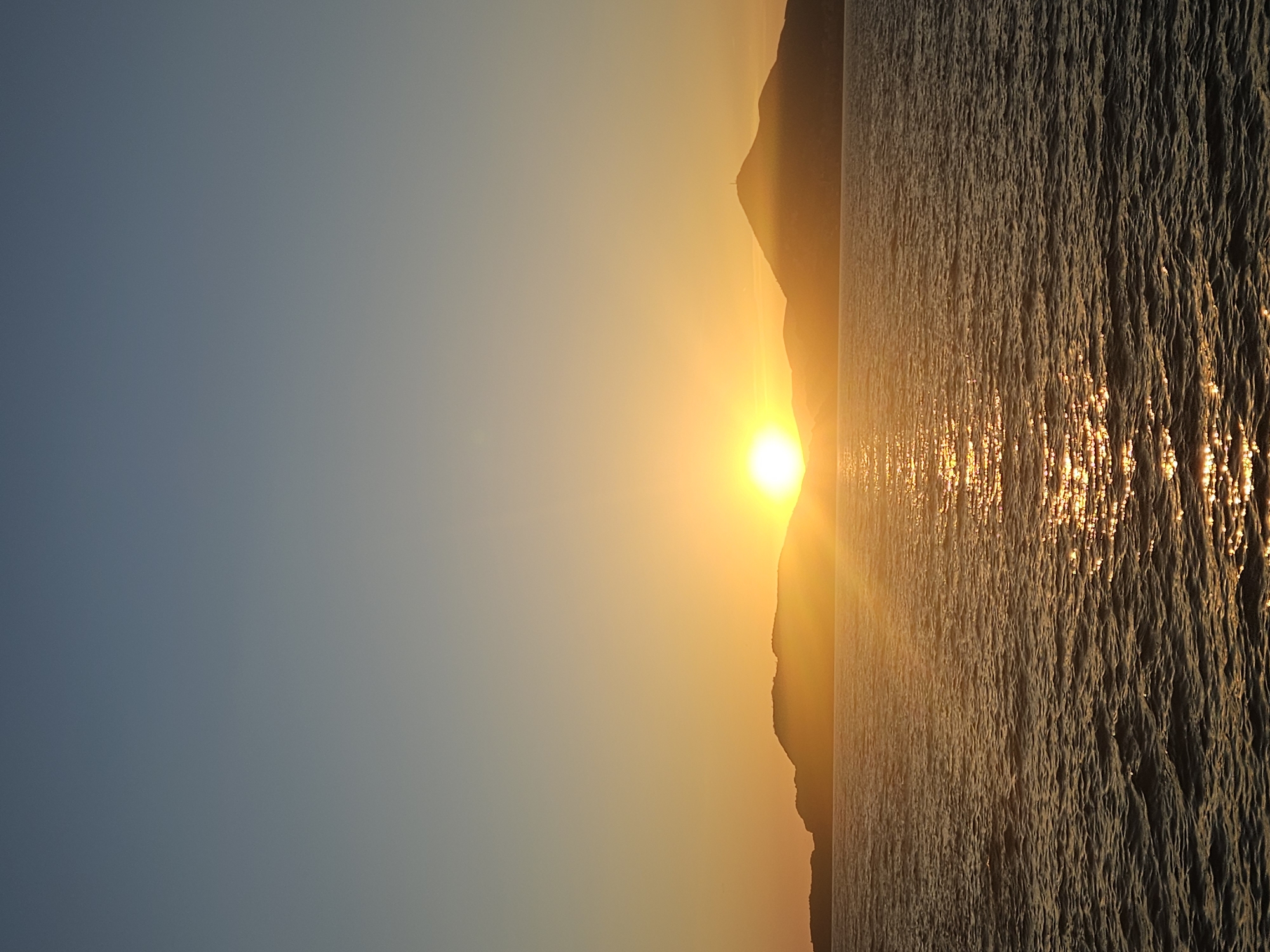 A serene landscape captured during either sunrise or sunset depicts a vibrant interplay of light and natural elements. A large body of water, likely an ocean, lake, or bay, dominates the lower and right portions of the frame, its surface textured with numerous small ripples and waves. A brilliant golden-yellow sun is positioned low above the horizon, casting a shimmering, elongated path of light across the water directly towards the viewer.
Silhouetted mountains form the horizon line, dark and imposing against the bright sky. The sun appears to be nestled between two peaks, with a prominent, jagged mountain rising to its right and a slightly lower, more rounded ridge to its left.
The sky transitions from a soft, clear blue-grey on the left to a warm, glowing golden-orange around the sun, indicating the early morning or late evening hours. There are no clouds visible. The scene is devoid of any people, animals, or man-made objects, focusing entirely on the raw beauty of the natural environment. No text is visible in the image.