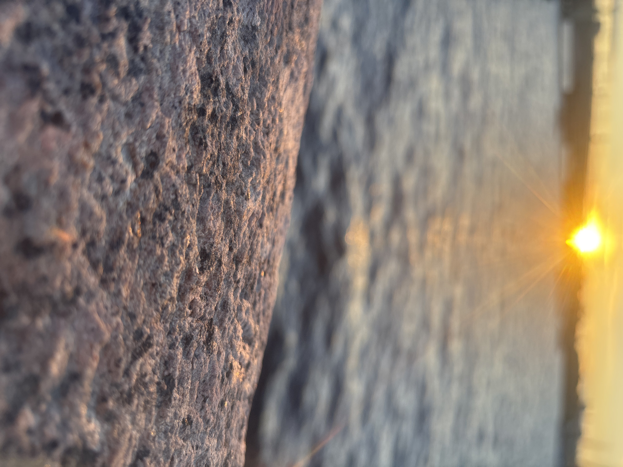 The image depicts a close-up, low-angle view of a rocky surface in the foreground, bathed in the warm, golden light of a sunset. The water of the Gulf of Finland is visible in the mid-ground, with gentle ripples reflecting the sunset's glow. In the far distance, a hazy silhouette of a bridge and possibly buildings along the St. Petersburg skyline can be discerned against the bright sun. The sky transitions from a soft yellow near the sun to a pale orange and then a muted blue. No people are visible in the image. The scene appears to be captured from the shore or a pier along the Gulf of Finland in Saint Petersburg, Russia, likely during the evening.