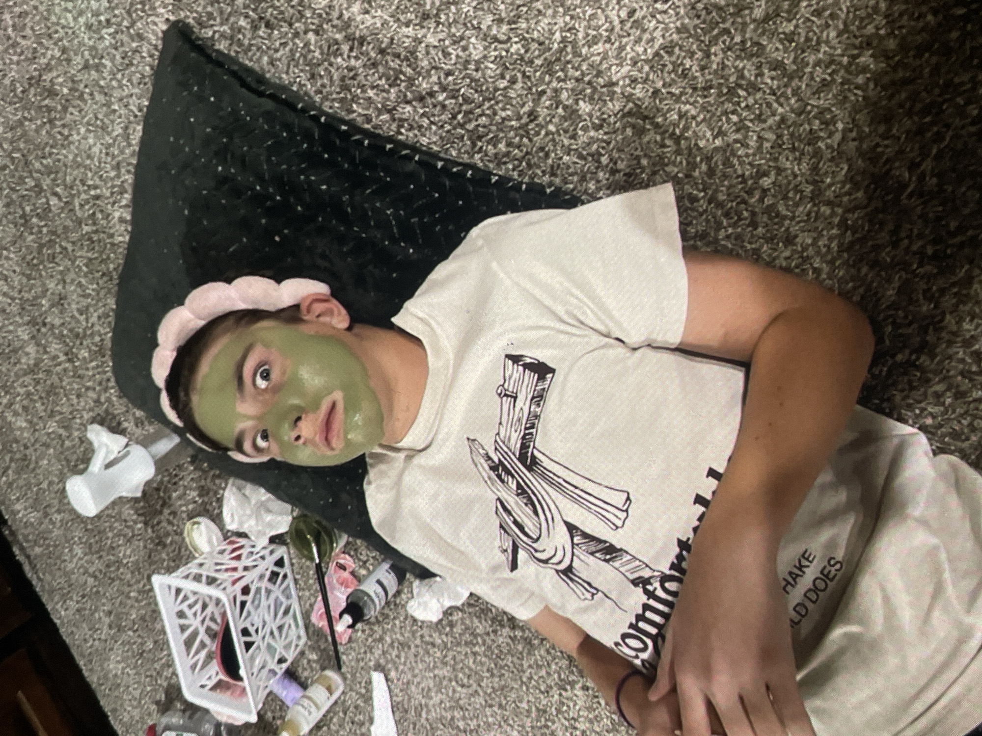 The image depicts a young person lying on a gray carpeted floor, resting on a black cushioned mat. The person is wearing a white t-shirt with a graphic design of a cross and cloth. A pink, puffy headband is worn across their forehead, and a green facial mask covers their face. Their eyes are wide open, and their mouth is slightly ajar, suggesting a surprised or playful expression. Scattered around the person are various personal care items, including bottles, a white basket, and what appears to be a brush and a small bowl. The overall scene suggests a relaxed or humorous moment, possibly during a skincare routine. The location is identified as Harmanli, Bulgaria.