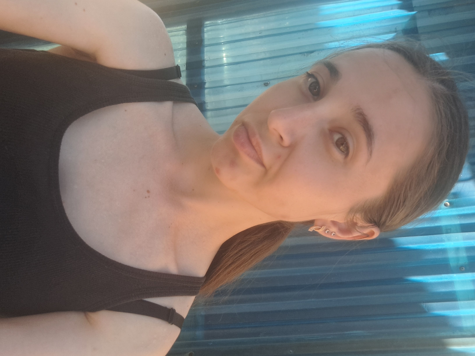 A young woman with light brown hair pulled back into a ponytail is posing for a selfie outdoors, against a blue, corrugated metal background. She is wearing a dark brown tank top. Her left ear has two small stud earrings and a larger, gold-colored earring. She has a light complexion and appears to be looking slightly off-camera. There is a faint red mark on her chin. The lighting suggests it is daytime, possibly late afternoon due to the warm, golden hue on her skin. The image was captured in Vypasne, Ukraine.