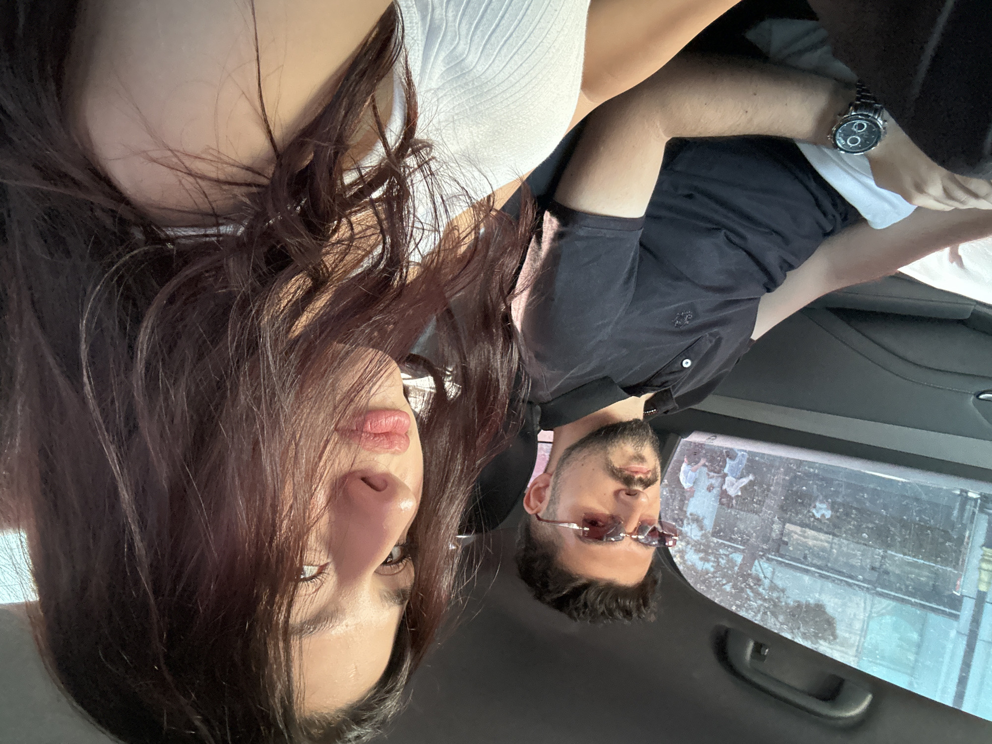 Two individuals, a male and a female, are seated in the interior of a vehicle, appearing to be in the city of Baku, Azerbaijan. The male, on the left, has dark hair and a beard, wearing sunglasses with pink lenses and a black polo shirt. He is looking forward. The female is in the foreground on the right, with long, dark hair and is looking towards the camera. She is wearing a white ribbed tank top. The background through the car window shows a street scene with trees and people, indicative of an urban environment. The male is wearing a wristwatch on his left arm. The vehicle's interior is visible, with dark-colored seats and a handle on the roof. The lighting suggests it is daytime.