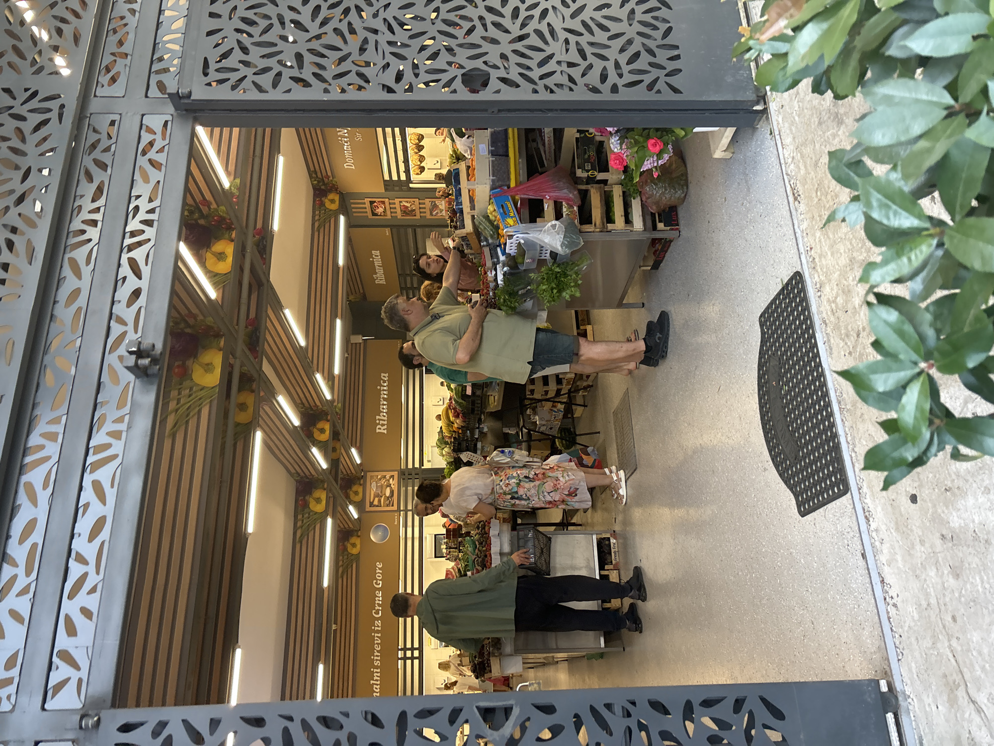 The image shows a fruit and vegetable market, likely located in Budva, Montenegro, as suggested by the text in the background. The market appears to be an indoor or covered establishment with a modern design featuring metal grating at the entrance and decorative elements on the ceiling.
Several people are visible, interacting with vendors and browsing produce. A man in a green jacket and black pants is standing near a stall, holding a basket. A woman in a floral dress is speaking to a man who is tending to the produce. Another woman is seen behind a counter, possibly a vendor. The stalls are laden with a variety of fresh fruits and vegetables, including what appear to be eggplants, berries, and other produce displayed in wooden crates. Some flowers are also visible, adding color to the scene. The overall atmosphere suggests a bustling marketplace.