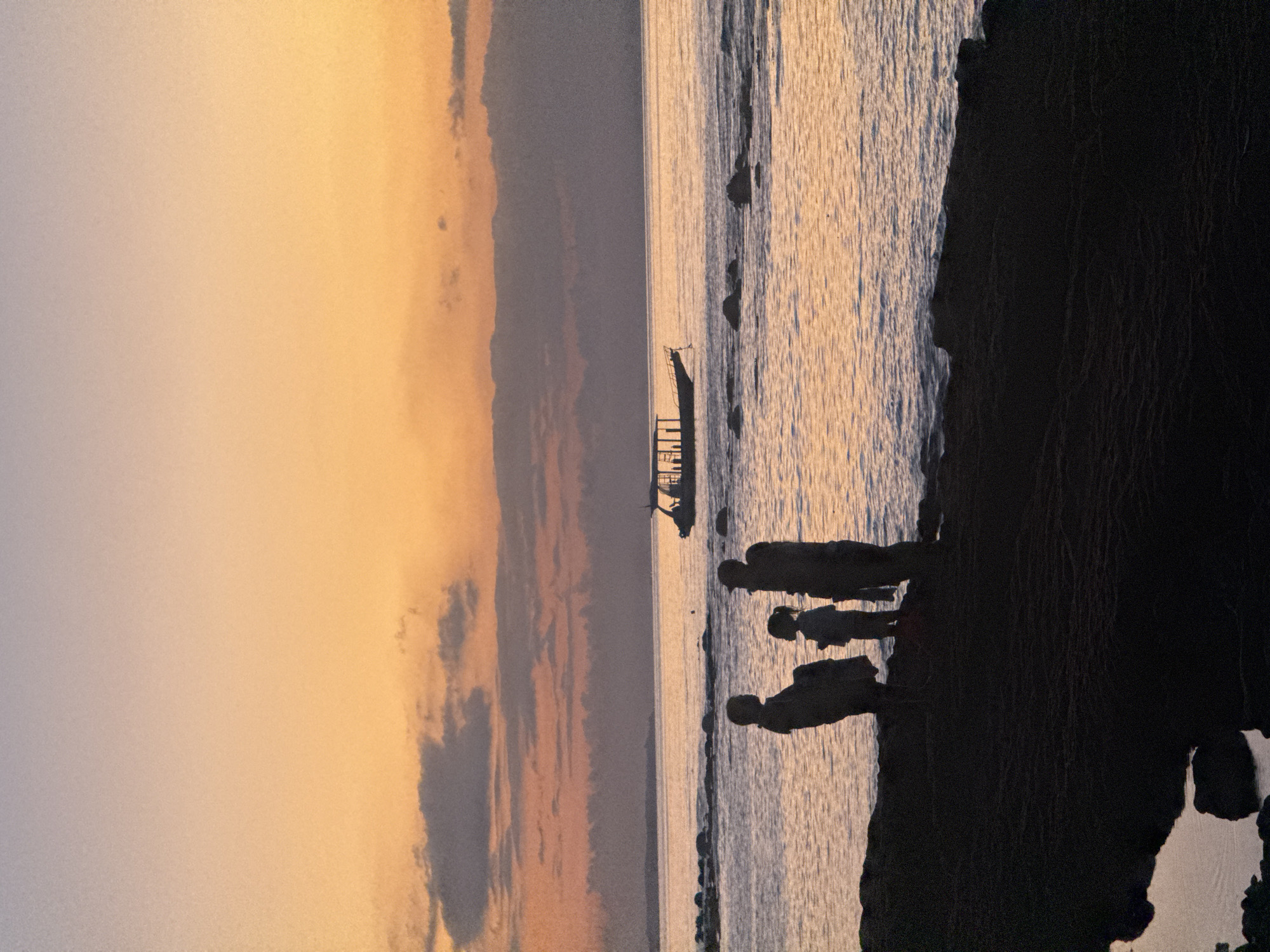 A family of three, silhouetted against a vibrant sunset, stands on a rocky shoreline in Nusa Penida, Indonesia. The sky is a gradient of soft oranges and purples, with a few scattered clouds adding texture. In the distance, a long boat with a canopy sails across the calm sea. The figures, two adults and a child, are positioned near the water's edge, their forms dark against the glowing horizon. The foreground is dominated by the dark, rugged terrain of the coast, with small pools of water reflecting the twilight sky.
