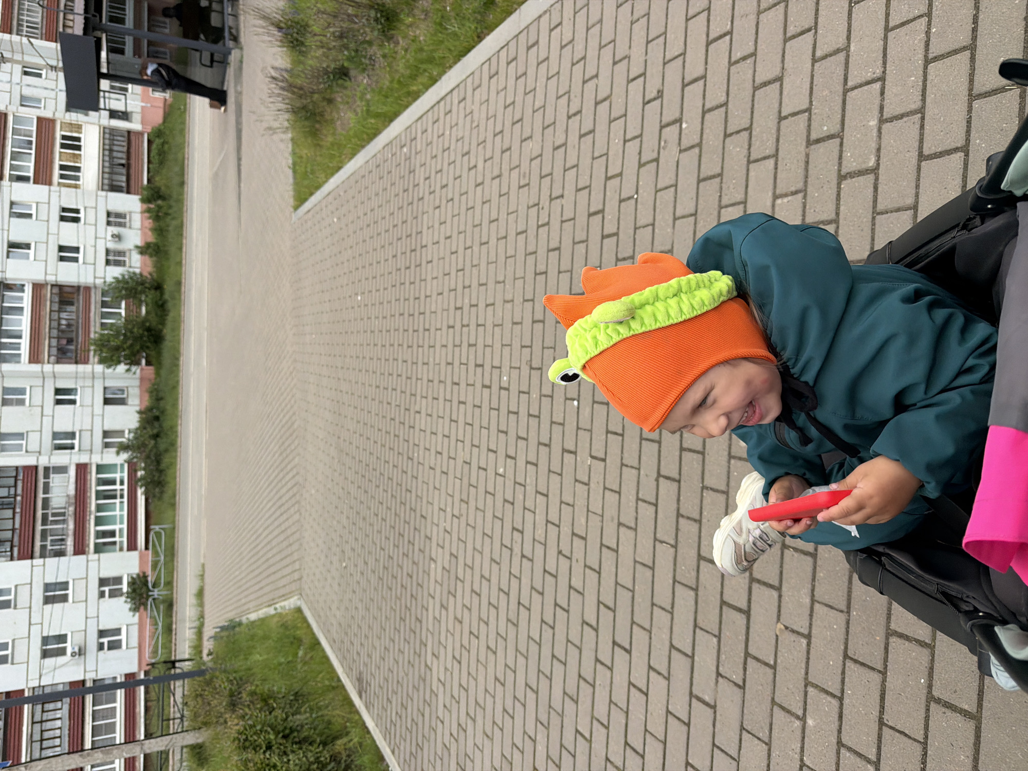 The image captures an outdoor daytime scene in Chaikovsky, Russia, featuring a young child in a stroller on a paved walkway, with a street, bus stop, and multi-story residential building in the background. In the foreground, slightly to the right, a child, estimated to be between two and four years old, is seated in a black and grey stroller. The child is wearing an orange knitted hat adorned with a bright green, plush, frog-like appliqué featuring two googly eyes. They are also wearing a dark teal or dark green hooded jacket and light-colored sneakers, with one foot slightly raised. The child is smiling broadly, with visible teeth and a dimple, and is looking towards the viewer's left. Both hands are holding a bright red, rectangular object, approximately 10-12 cm in length, which appears to be a toy or personal item. A bright pink fabric element is visible within the lower left compartment of the stroller. The ground beneath the stroller and extending into the midground is a paved pedestrian walkway constructed from light grey rectangular pavers arranged in a running bond pattern. To the left of the walkway, a subtle grassy incline features sparse green vegetation and small bushes. A light-colored curb delineates the edge of the path. Further left, a dark-colored utility pole is partially visible, and beyond it, a low, two-section metal barrier with horizontal bars stands behind the greenery. In the midground, an asphalt road surface is visible, separating the pedestrian area from the background structures. To the right of the road, a modern-style black bus stop shelter with a solid roof and a clear or glass rear panel is present. Two individuals are at the bus stop: one person in dark clothing, possibly a black jacket, is leaning against the right side of the shelter, facing away from the viewer; another individual is seated on the bench inside the shelter, largely obscured. The background is dominated by a multi-story residential apartment block, characteristic of urban architecture in Chaikovsky, Russia. The building has a light grey or off-white facade with numerous rectangular windows, mostly framed in white. Several balconies are visible, featuring dark red or brown bases; some are open, while others are enclosed with glass, including a distinctive ground-floor balcony on the far left with green-tinted windows. Small deciduous trees and low vegetation are sparsely planted in the green space between the road and the residential