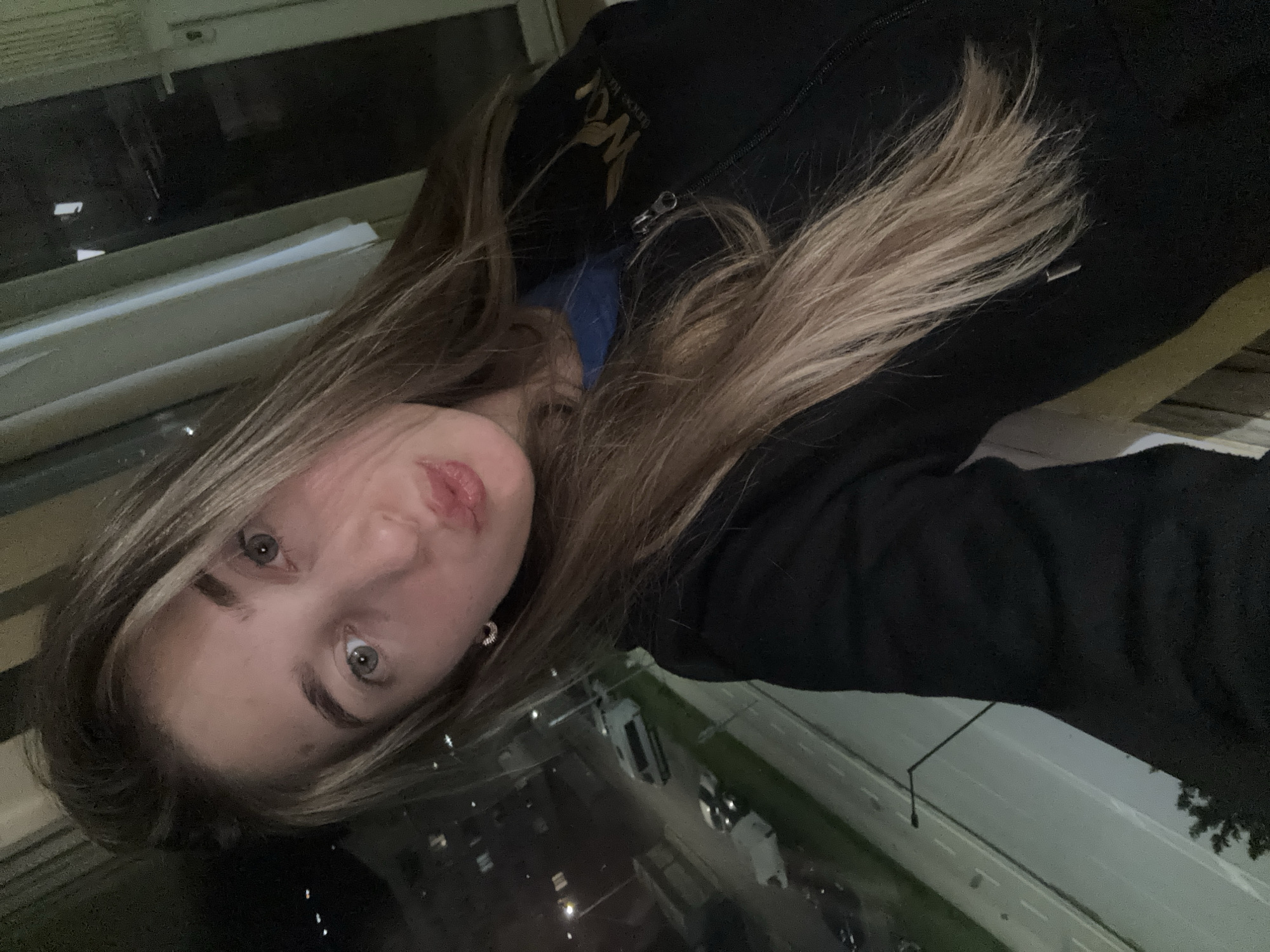 A young woman with long, blonde hair and light eyes is posing for a selfie. She is wearing a dark hooded sweatshirt with a logo on the upper left. She has fair skin and is wearing earrings. The background shows a nighttime urban scene, with street lights and vehicles visible through what appears to be a window. The city location is unidentified.