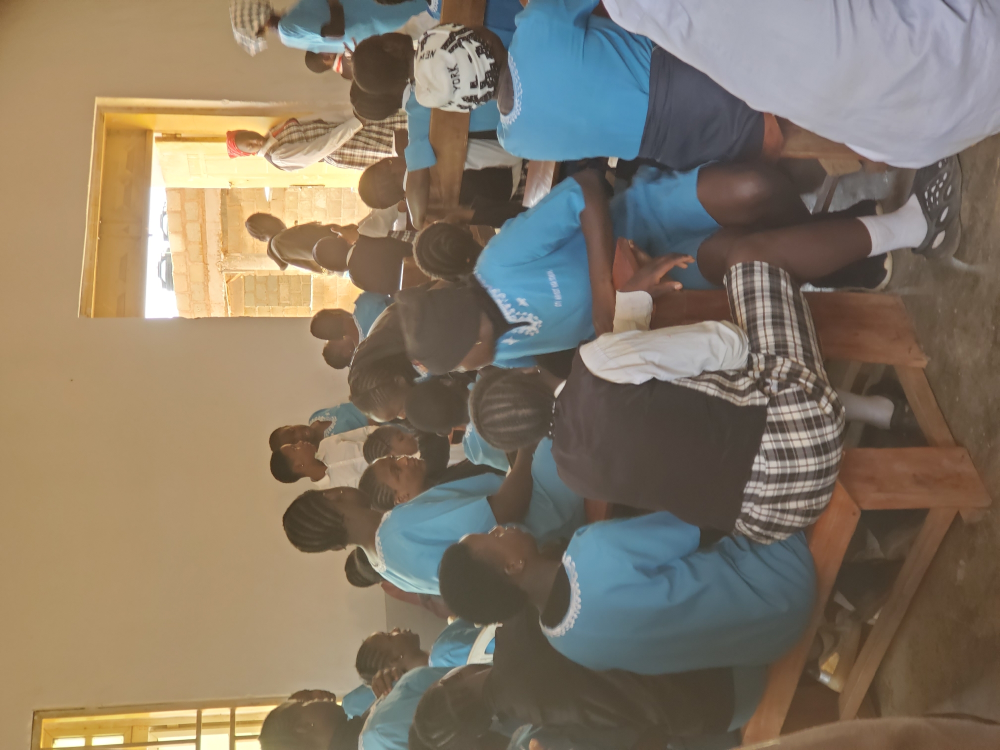 The image depicts a group of people, likely children and young adults, gathered indoors. They are seated on wooden benches, facing away from the camera, and appear to be engaged in a lesson or activity. Many are wearing matching light blue shirts, some with white trim around the neckline and sleeves. One individual in the foreground wears a white bandana with "NEW YORK" printed on it. Another person is wearing a plaid shirt.
The setting appears to be a simple room with a window on the left, through which natural light is streaming in, suggesting it is daytime. The walls are a light color, and the floor is visible in some areas, appearing to be concrete or dirt. The overall atmosphere seems calm and focused. There are no explicit indications of the weather or time of day beyond the natural light. The image does not convey strong emotions, but rather a sense of quiet engagement.
The visible text includes "NEW YORK" on a bandana. The cropped images provide closer views of some individuals' attire and the benches they are seated on, highlighting the details of their clothing and the wooden furniture. The context of Kuru Karama, Nigeria, suggests a community or educational setting.