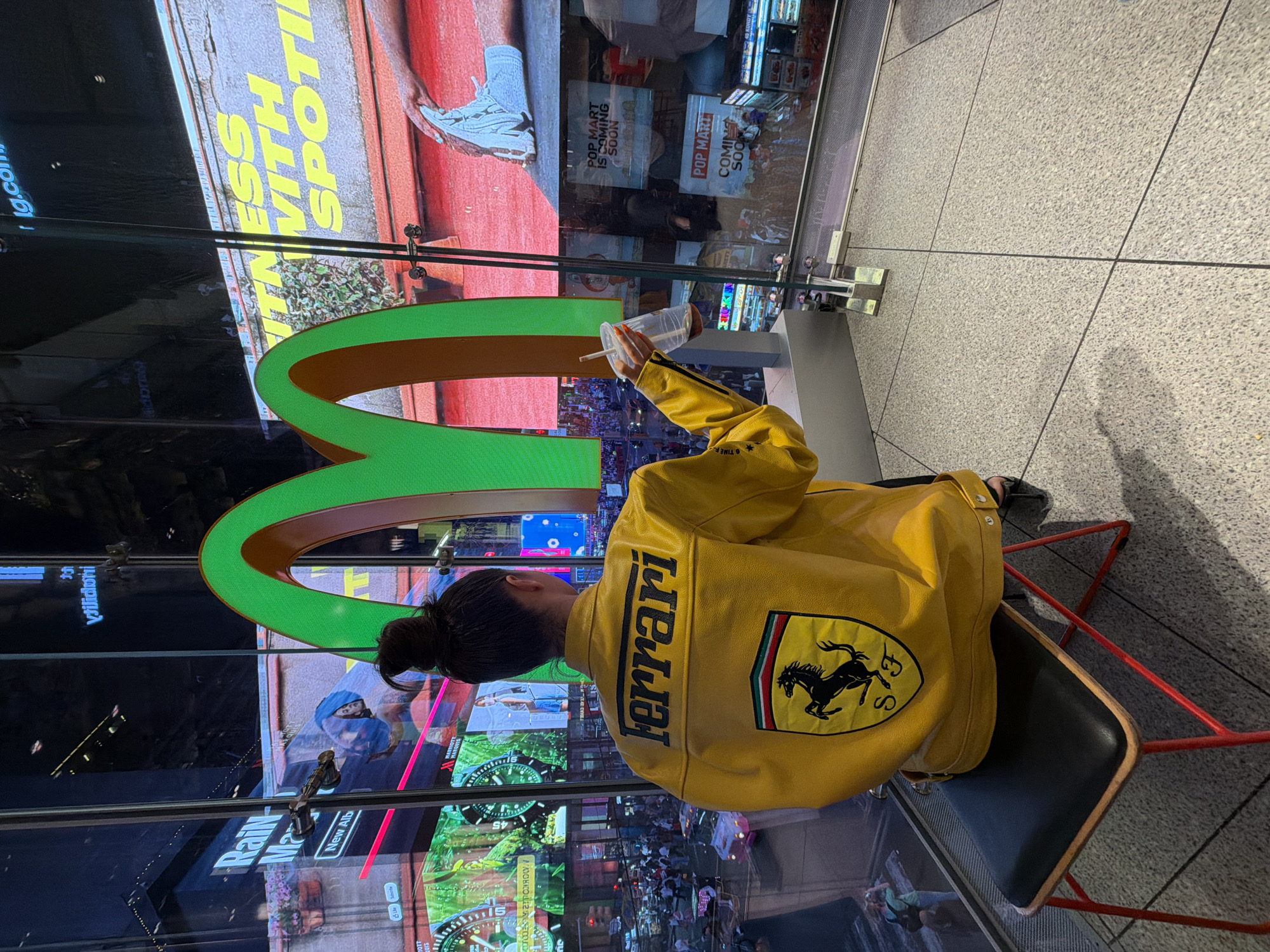A person is seated on a chair holding a McDonald's beverage cup in front of a large display in Times Square, New York City, New York, United States of America. The person is wearing a bright yellow jacket with "Ferrari" emblazoned on the back, along with the Ferrari emblem, and has dark hair pulled up in a bun. The large display has an advertisement for "Fitness With Spotify" and "Pop Mart Is Coming Soon". The display also shows a variety of other images and advertisements, including a McDonald's logo. The chair the person is sitting on has a black seat and a red metal frame. The floor is tiled.