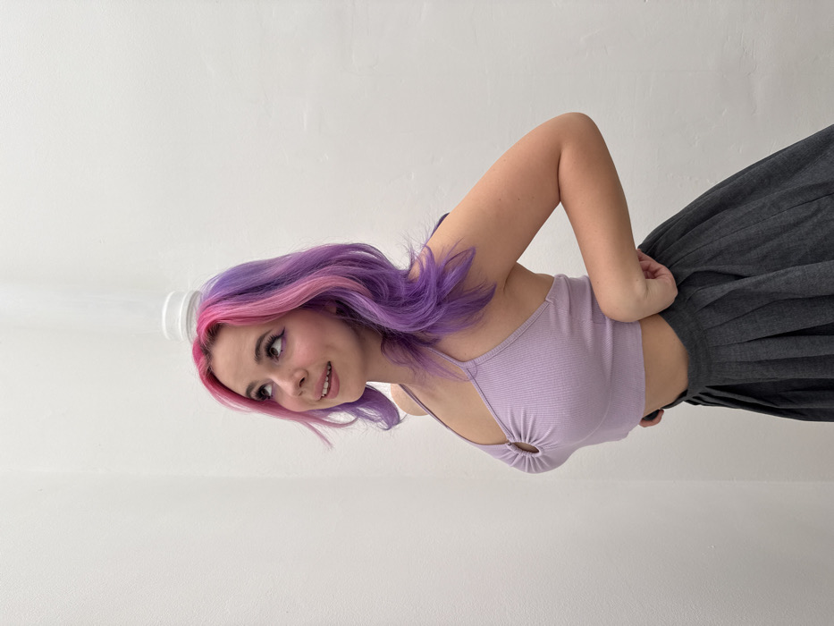 A young adult female is depicted in a half-body portrait against a plain white textured wall, described as being in the City of Zagreb, Croatia. Her shoulder-length hair features a split dye of pink on the viewer's left and purple on the viewer's right. She looks upwards and to her left with a subtle smile, her lips slightly parted. A white, inverted disposable cup is balanced precisely on the top of her head. She wears a light purple ribbed halter-neck crop top with a circular ring detail at the center of the chest and a dark grey pleated skirt. Her right hand is positioned at her waist, appearing to adjust the skirt fabric.