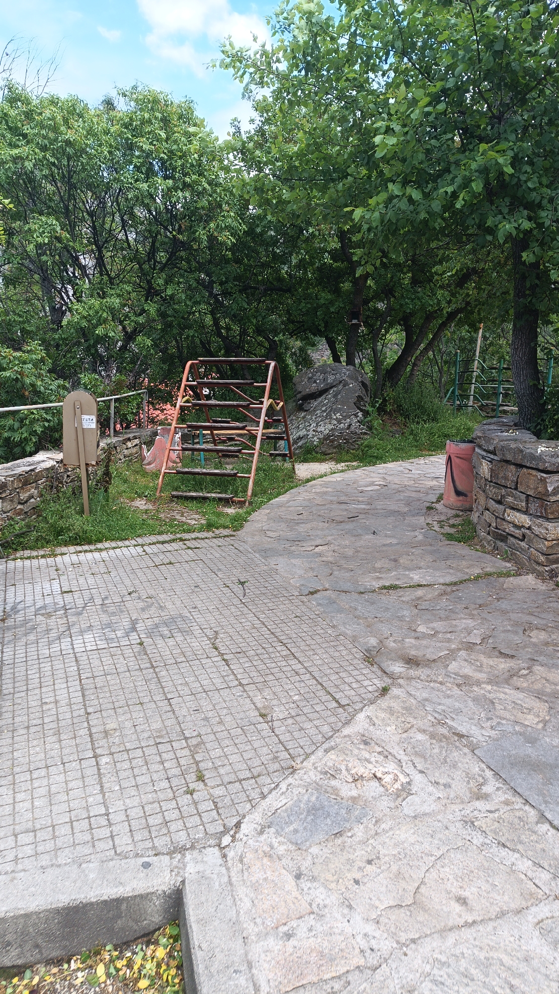 The image shows a park in what is likely Lyaskovo, Bulgaria, given the prompt's request. The ground surface is a mix of tiled and stone pathways. An old, partially dilapidated playground structure is present. Trees and other greenery surround the area. A stone wall and a metal sign are visible. A red receptacle sits to the side of the stone pathway. No people are visible in the image.