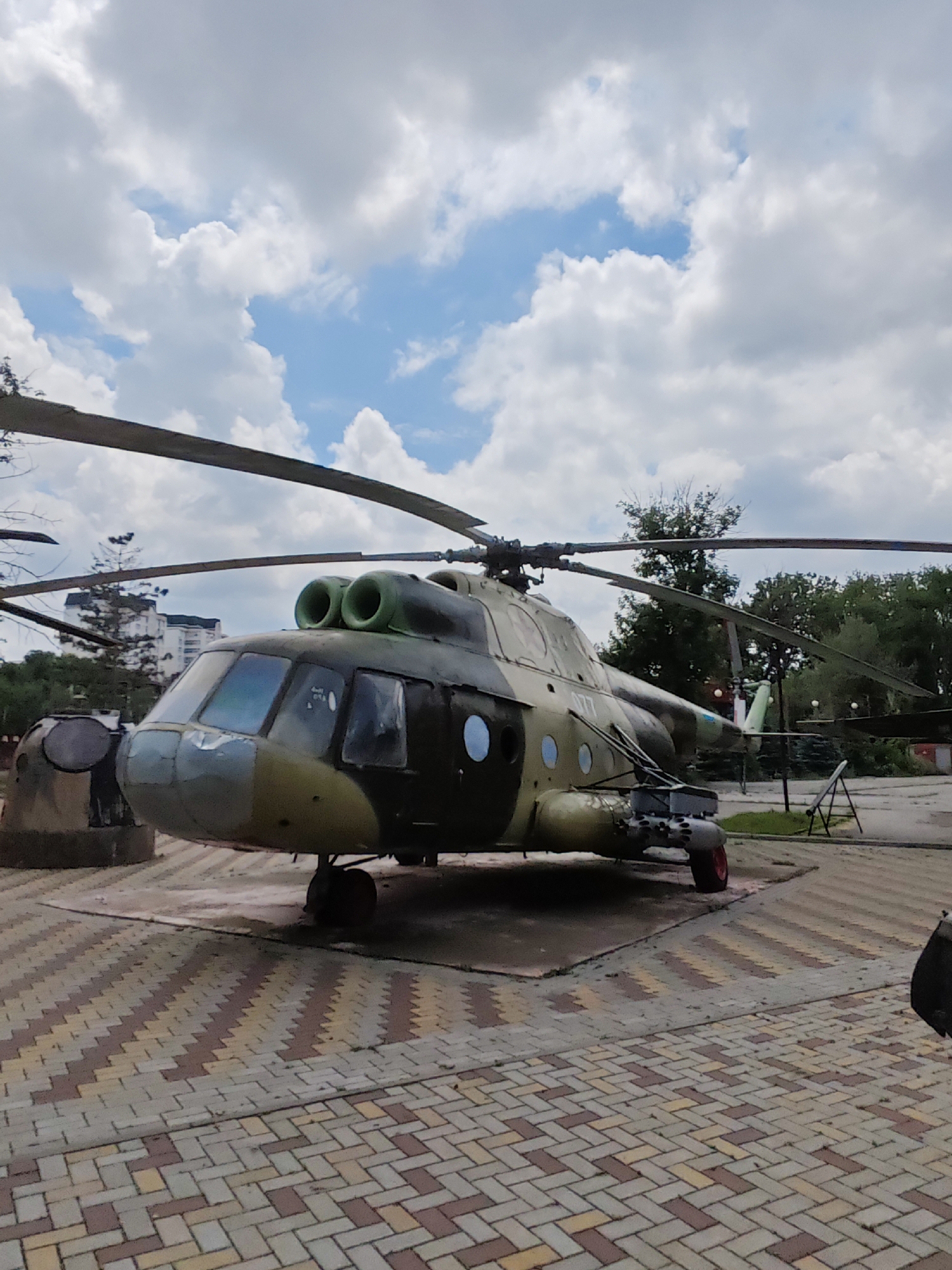 An olive-drab military helicopter, likely a Mi-8 variant due to its distinctive twin-engine nacelles and overall shape, is displayed outdoors on a paved area in Cherkessk, Russia. The helicopter is angled slightly towards the viewer's left and its rotor blades are stationary. The paving consists of interlocking bricks in shades of red, yellow, and gray, arranged in a herringbone pattern. In the background, lush green trees frame the scene, and a multi-story building, typical of urban Soviet-era architecture, is visible behind them. The sky above is a vibrant blue, dotted with fluffy white clouds. The helicopter appears to be a static exhibit, possibly in a park or museum, given its placement and the surrounding environment.