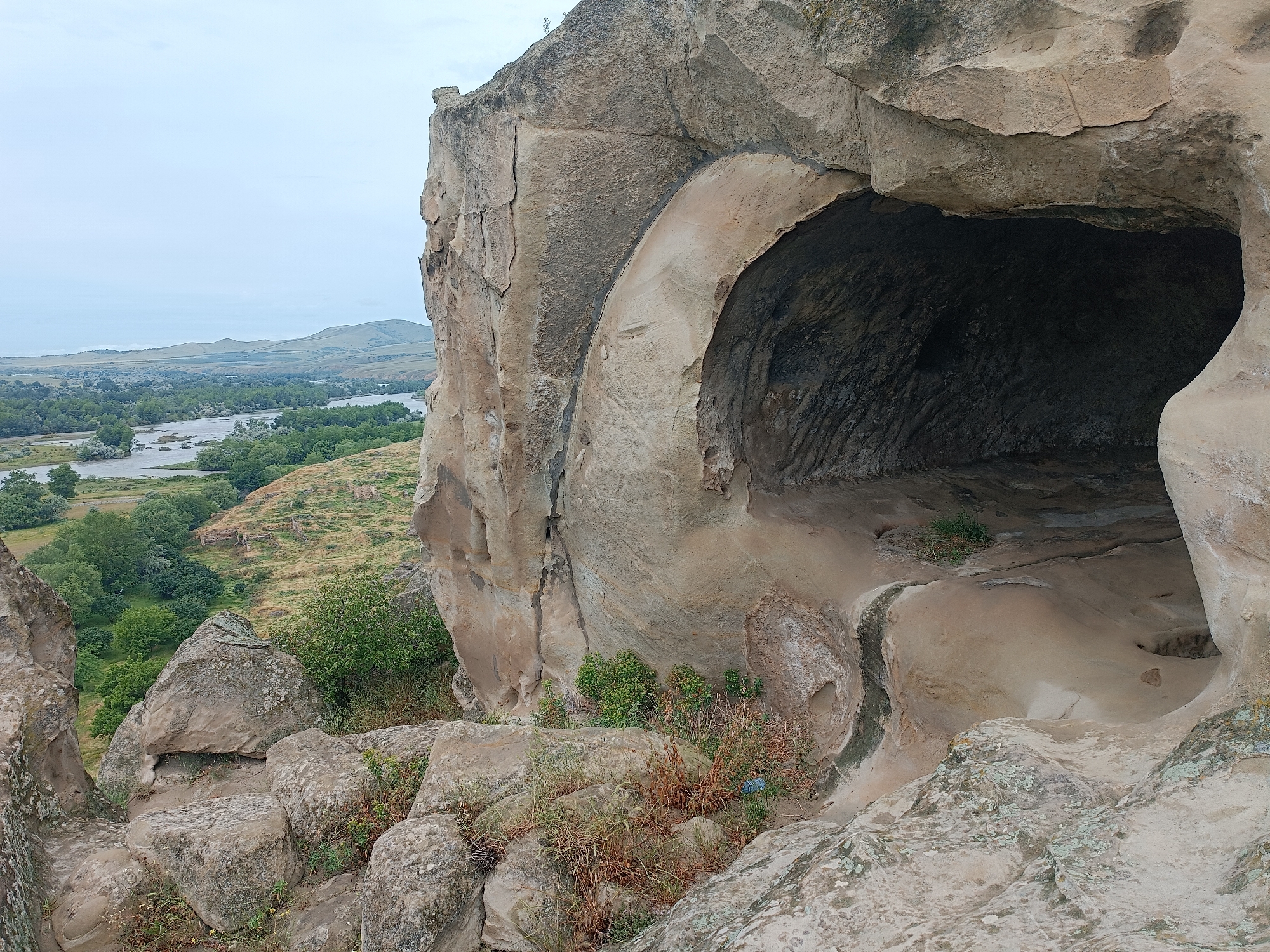 The image shows a view from within a cave-like opening carved into a large rock formation. The opening is arched and the interior is dark and textured. Outside the opening, in the foreground, are large, weathered rocks with sparse vegetation. In the mid-ground, a river winds through a valley with green trees and rolling hills. In the background, distant hills meet a hazy sky. The scene is located in Uplistsikhe, Georgia, an ancient rock-hewn town.