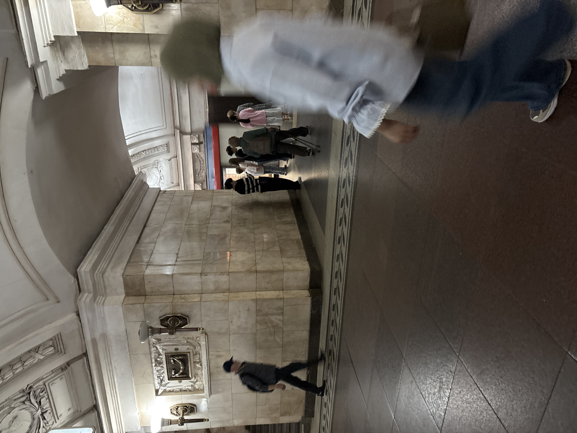 The image depicts a scene in a Moscow Metro station. In the foreground, a person in a blue shirt and jeans is walking away from the camera, their face blurred with motion. Behind them, a group of people are standing in a line, possibly waiting for a train. One person in the group is wearing a green jacket and carrying a backpack, while another wears a pink shirt. To the left, a man in a cap and backpack is walking towards the left side of the frame. The station features ornate architecture with marble tile walls, decorative ceiling moldings, and antique-style lighting fixtures. The floor is tiled in a dark, glossy material with a patterned border. The overall impression is of a busy, public transportation hub with a classic, somewhat aged aesthetic.