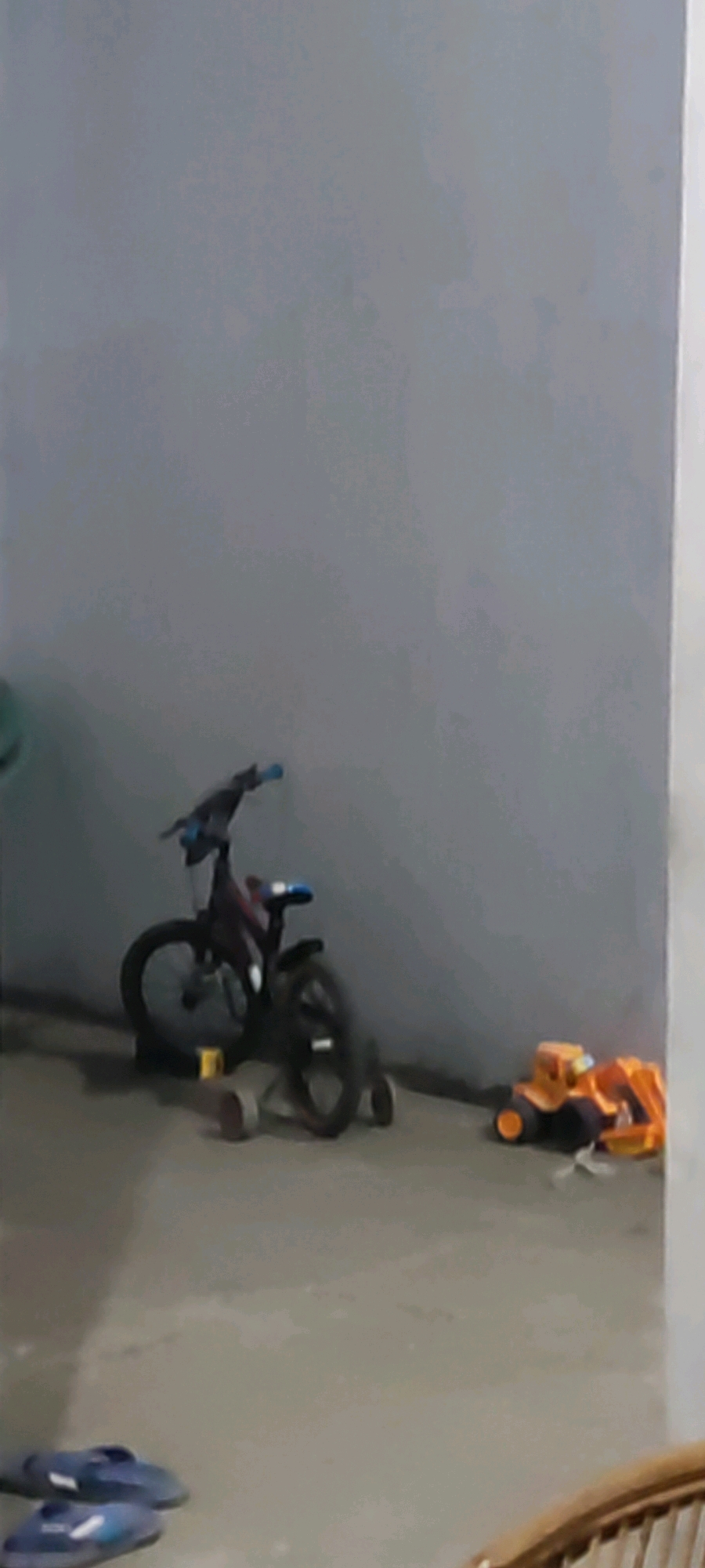 The image depicts a dimly lit indoor scene, possibly a garage or patio, located in Karangwangun, Indonesia. The background is a plain, textured gray wall. In the mid-ground, a dark-colored bicycle with blue handlebars is leaning against the wall. To the right of the bicycle, a yellow and orange toy construction vehicle, resembling an excavator, is present on the concrete floor. In the foreground, two dark blue slippers are lying on the floor. The lighting is low, creating shadows and reducing the clarity of details.