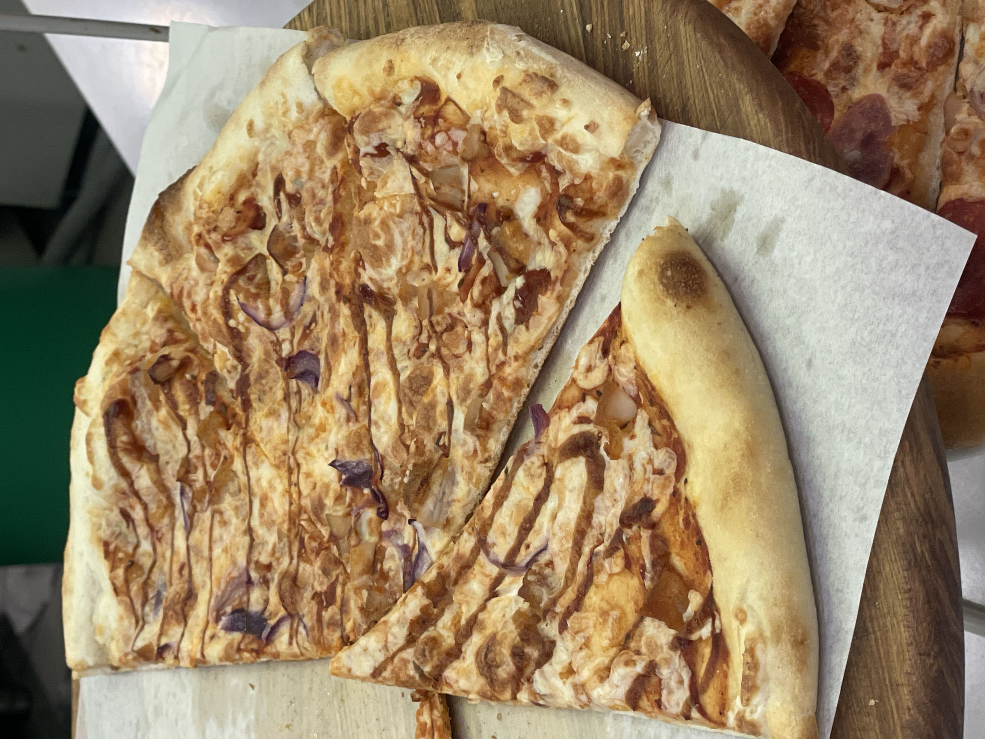 A close-up, eye-level, medium shot shows two slices of pizza on a wooden board. The pizza appears to have a tomato sauce base, cheese, red onions, and strips of chicken. The pizza is on a white paper liner. The crust is golden brown. In the background, a green surface is visible. The lighting is artificial. The scene is indoors, possibly in a restaurant or home kitchen in Sochi, Russia. No people are visible in the image.