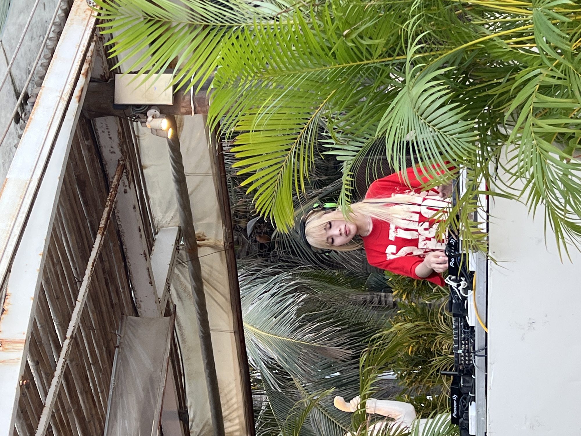 The image shows a woman with blonde hair and green highlights wearing a red long-sleeved shirt, standing behind a Pioneer DJ setup. The setting appears to be outdoors, framed by lush tropical vegetation, likely palm trees. A section of metal architecture is visible on the left. Based on the presence of the Pioneer DJ equipment and the general tropical environment, the location is possibly an outdoor event space or bar in Nha Trang, Vietnam, as the city is known for its coastal setting and vibrant nightlife. However, without further details, the specific location within Nha Trang is not identifiable.