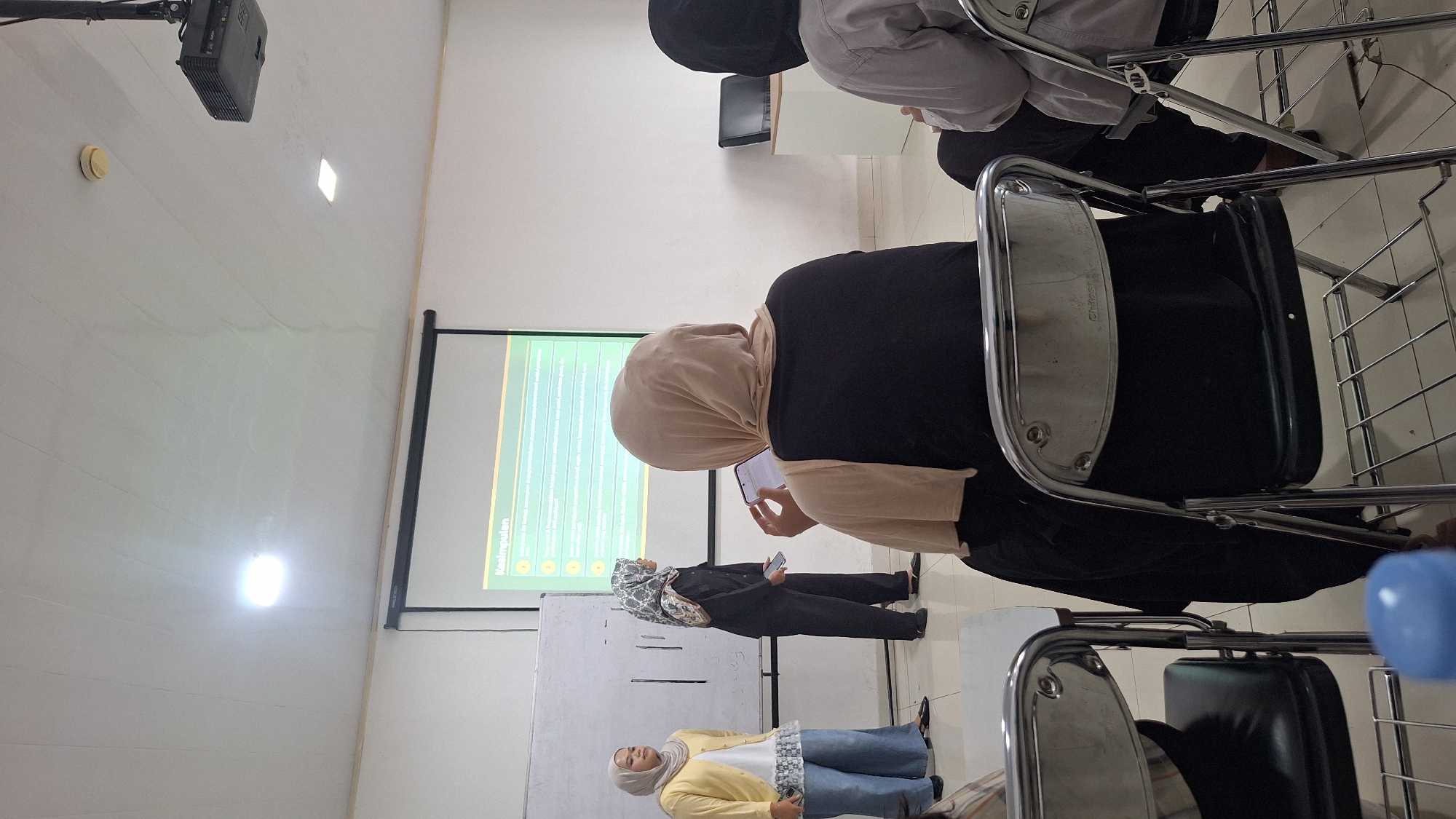 A vertically oriented photograph captures a room filled with people. The foreground features a row of metal chairs. Several people are seated, and others are standing near a whiteboard and a screen displaying text and bullet points. All the women are wearing headscarves. One person is speaking, while another is looking at a phone. The room is well-lit and appears to be an indoor educational or presentation setting. The location is Sukaharja, Indonesia.