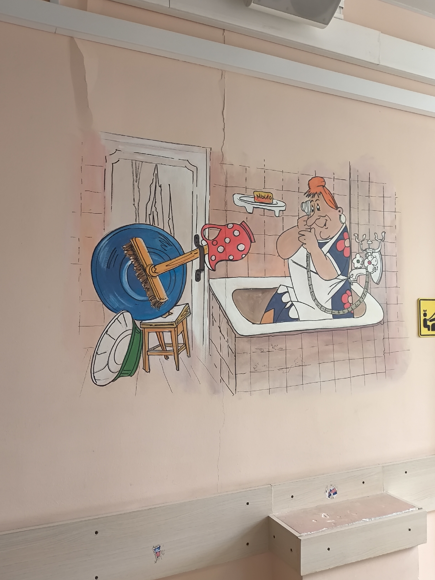 The image depicts a cartoon mural on a light pink wall. The mural depicts a bathroom scene with a woman in a bathtub talking on an old-fashioned telephone. To the left, there is a drawing of a door, a stool, a washbasin, and a brush. The style of the mural is simplistic, with bold outlines and bright colors. There's a crack in the wall above the mural. The lower part of the wall has a light-colored shelf-like structure attached to it. Small stickers are visible on the wall near the bottom and on the shelf-like structure. There's a yellow sign with a pictograph to the right of the mural.
While the image is identifiable as being in Russia because the word "Mыло" (soap in Russian) is visible on the mural, there is no indication of the city of Khimki. I am unable to find evidence supporting this claim. The location is not identifiable from this image.