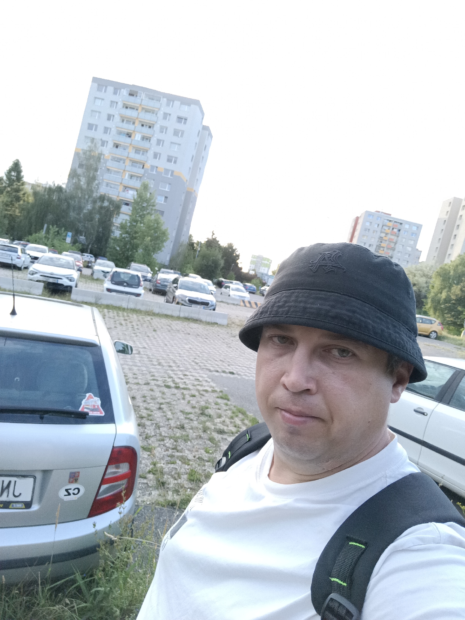 A selfie taken by a man wearing a black bucket hat and a white t-shirt, with a backpack over his shoulders, captures a parking lot scene in a residential area of Prague, Czechia. The background reveals several apartment buildings, typical of Prague's suburban districts, with a scattering of parked cars. The man is positioned in the foreground, looking directly at the camera, while a silver car is partially visible to his left. The environment suggests an outdoor setting, possibly during the late afternoon or early evening, with the sky appearing bright.