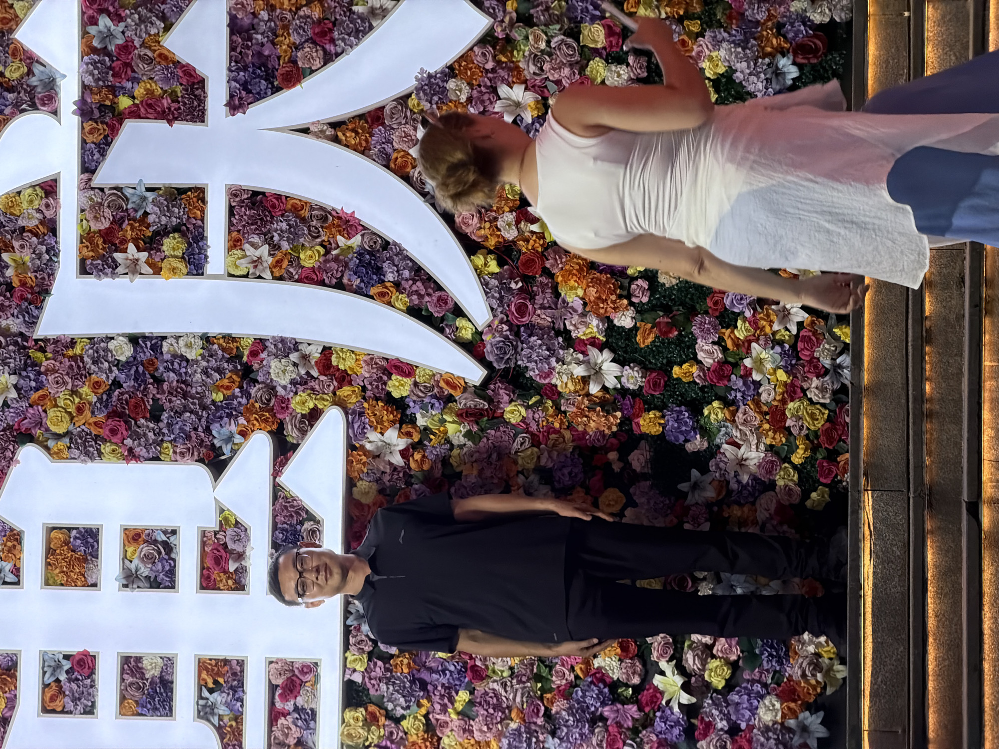 The image shows two people standing in front of a large floral display that is part of a building facade in Nan'an District, China. The floral display is composed of numerous artificial flowers in vibrant colors, predominantly shades of pink, purple, yellow, and orange. Large white Chinese characters are mounted on the floral wall, illuminated from behind.
On the left, a man wearing glasses, a black polo shirt, and black pants stands facing the camera. He is positioned on stone steps that are lit from beneath with warm, orange lights.
On the right, a woman with her back to the camera is seen in profile, looking towards the right. She is wearing a white sleeveless top and a light-colored, flowing garment over blue pants. She is also standing on the illuminated steps.
The overall impression is that of a celebratory or decorative backdrop, likely for an event or establishment. The lighting suggests it is evening or nighttime.