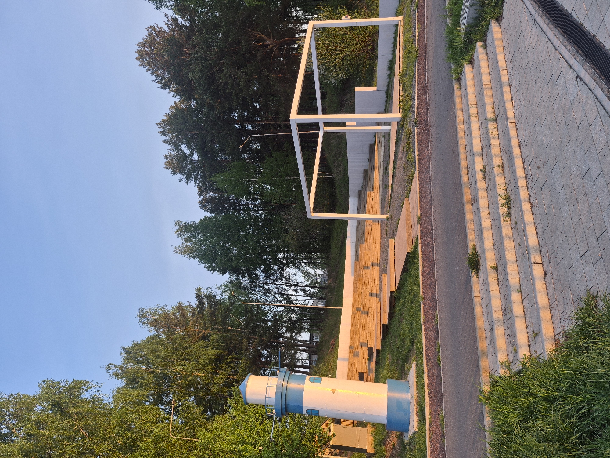 The image presents an outdoor scene in the city of Chaikovsky, Russia. A blue and white lighthouse replica stands on a concrete base near a paved walkway. Adjacent to the walkway are wooden structures or benches and a white, cube-like metal framework. Further back are trees and a blue sky. The environment is illuminated with the warm light of late afternoon or early morning. There are no people depicted in the image.