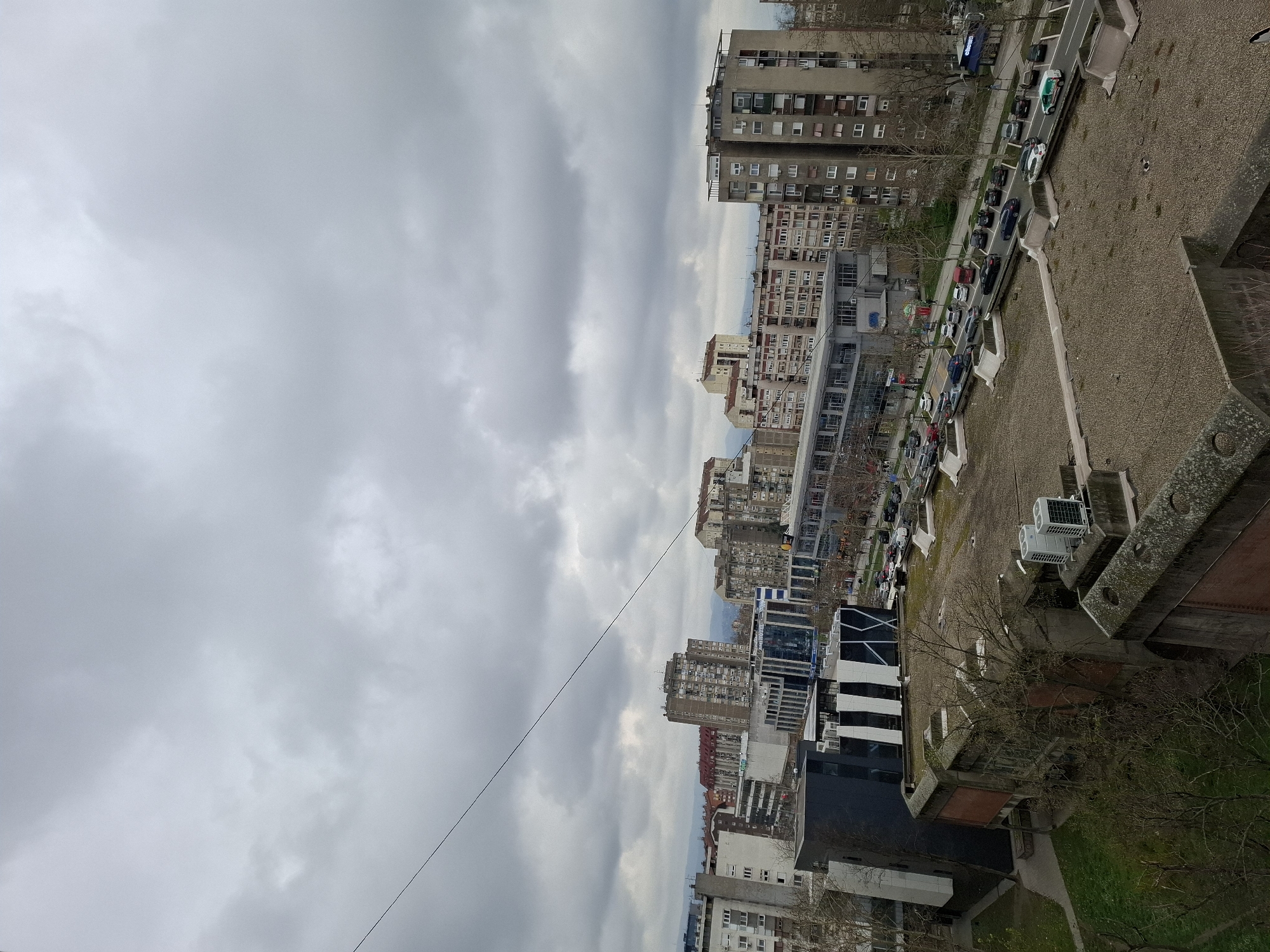 A high-angle view depicts a densely populated urban area in Novi Sad, Serbia, under an overcast sky. Rows of apartment buildings of varying heights and architectural styles dominate the scene, interspersed with commercial structures. Streets are lined with parked and moving vehicles, including cars and a green and white ambulance. The foreground includes a rooftop with air conditioning units and a partially visible green space with bare trees. A thin cable stretches across the sky, adding a linear element to the scene. The perspective suggests the image was taken from a tall building or elevated position.