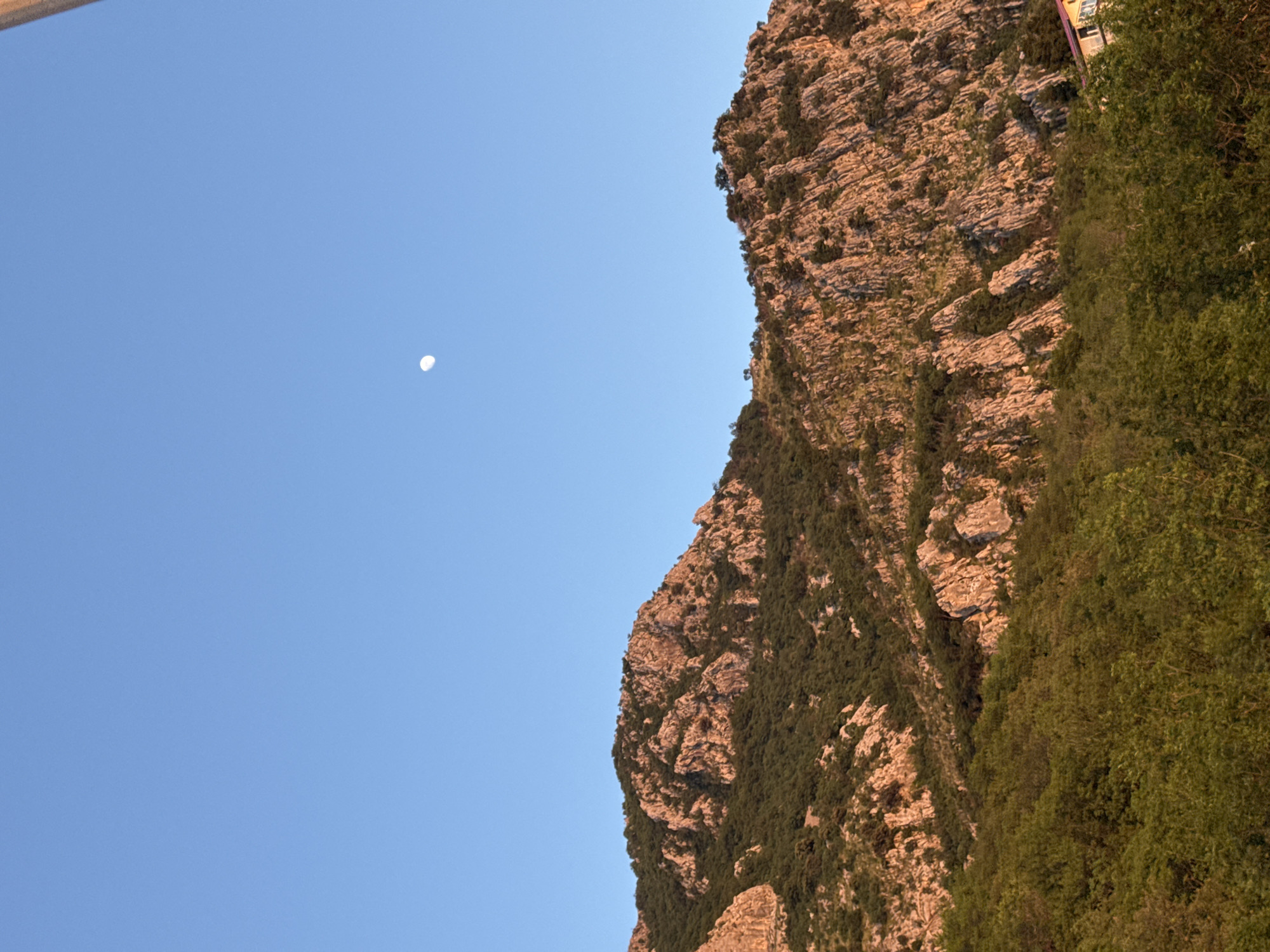 The image shows a partially visible rocky mountain face with sparse vegetation against a clear blue sky, with a crescent moon visible in the sky. The scene appears to be in natural light. The location is Tudorovići, Montenegro.