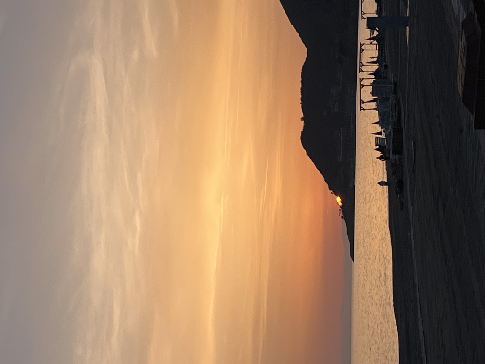 The image depicts a sunset over the Mediterranean Sea in Alanya, Turkey. The sun is setting behind a mountain, casting an orange and pink hue across the sky and reflecting on the water. A beach is visible in the foreground with lounge chairs and other beach equipment. The photo appears to have been taken from a slightly elevated position.
