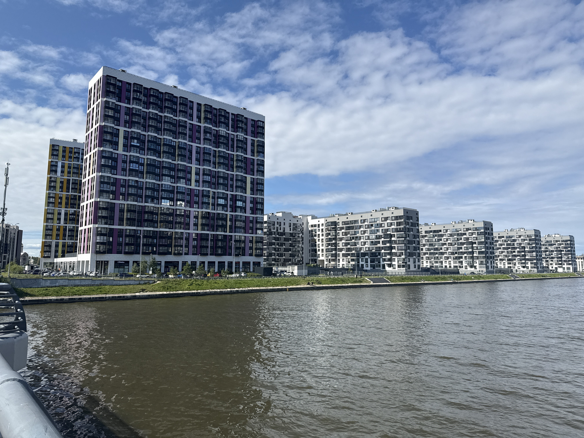 The image shows a waterfront view of a modern residential complex in Saint Petersburg, Russia, likely in the Primorsky district, based on the architectural style and proximity to the Gulf of Finland or a large canal. In the foreground, a portion of a grey railing and a concrete structure is visible on the left, suggesting the photo was taken from a bridge or embankment. The middle ground features a wide, dark body of water with ripples on its surface, reflecting the sky. Along the opposite bank, a line of multi-story apartment buildings stretches across the frame. The closest buildings on the left are taller and more colorful, with facades featuring alternating bands of purple, white, yellow, and dark grey, interspersed with numerous windows. Further to the right, the buildings become uniformly white and grey, with repetitive patterns of balconies and windows. A green strip of lawn and trees separates the buildings from the water, and a road with parked cars and a few pedestrians is visible in front of the buildings. The sky above is partly cloudy, with patches of blue sky and fluffy white clouds. A cell tower is visible on the far left, behind the first apartment building.
