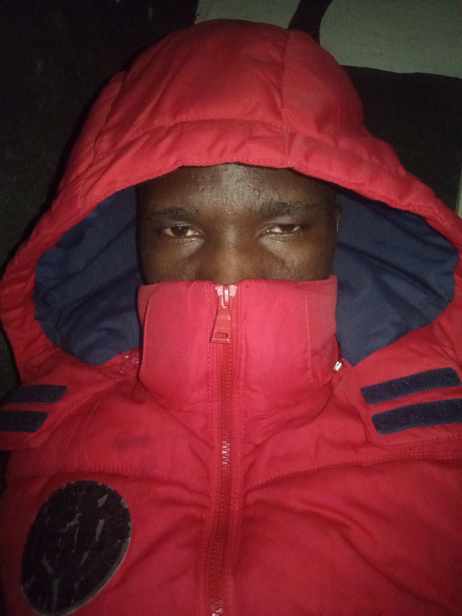A dark-skinned person is wearing a red hooded winter jacket with the hood up and partially zipped up, concealing the lower half of their face. The interior lining of the hood is navy blue. The jacket features dark rectangular patches on the shoulders and a dark, round patch on the lower left. The background appears dark and indistinct. The location is Unknown, Unknown.