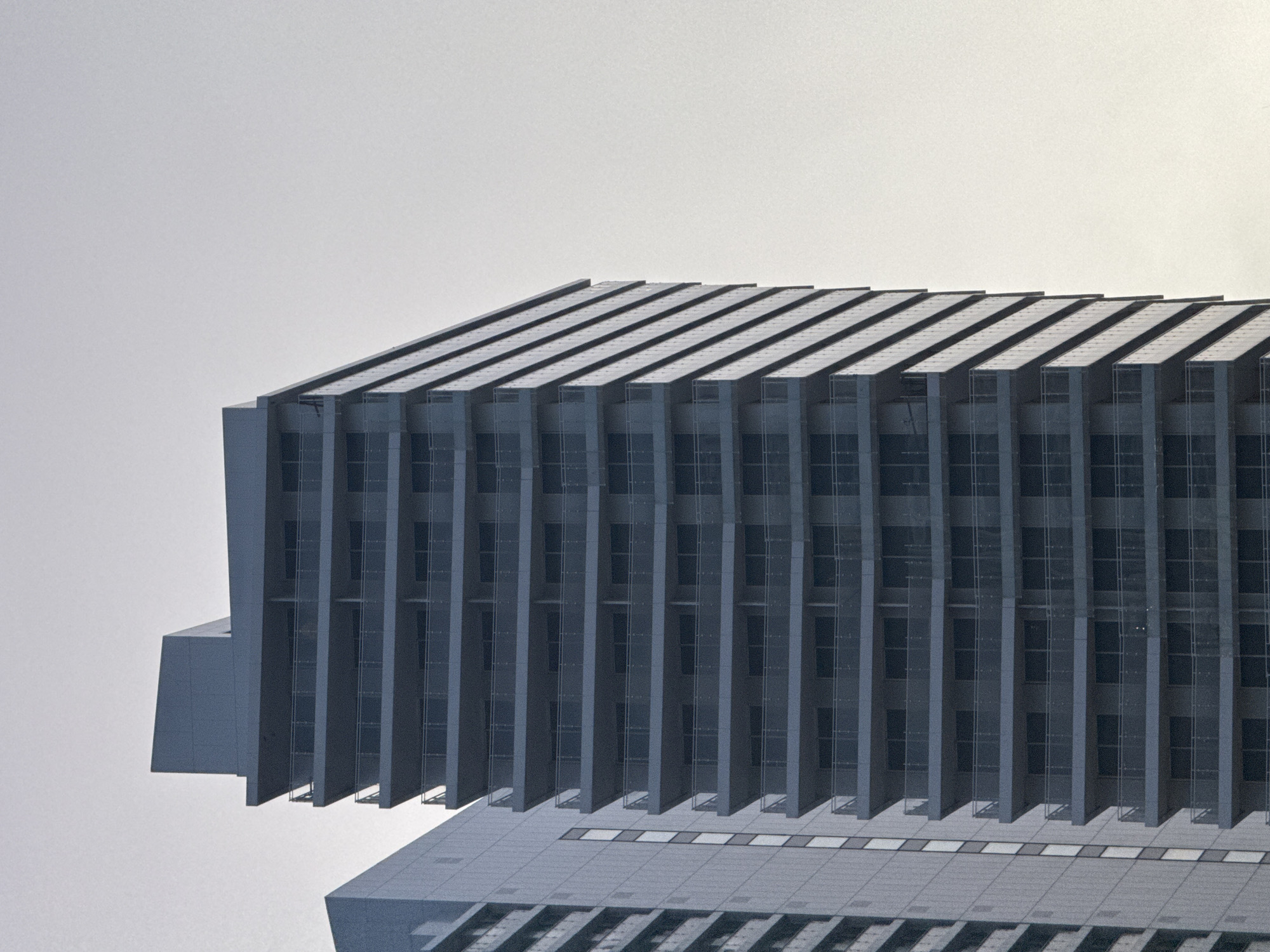 An eye-level, exterior shot shows the upper portion of a modern building in Bat Yam, Israel. The building features a striking, repetitive architectural design with long, dark gray or blue rectangular protrusions extending horizontally along the façade. These protrusions cast distinct shadows. The building appears to have multiple windows, seen through the gaps between the protrusions. The sky is a uniform, soft gray-white color, suggesting a cloudy or overcast day. The lower portion of the building is visible, displaying a rooftop or upper level with square tiles or panels.