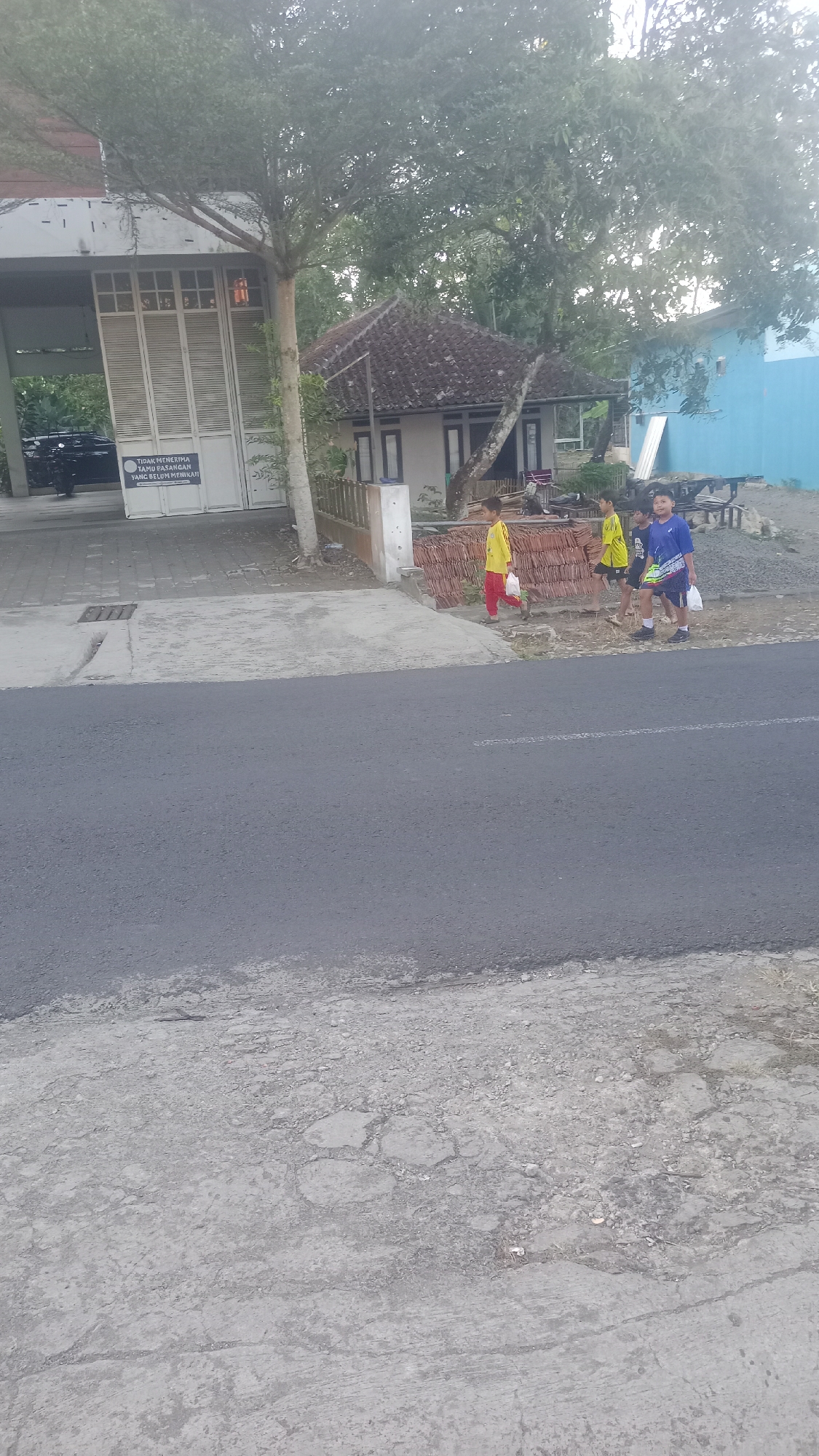 In Karyamandala, Indonesia, on a paved road, three young males walk alongside a dirt path. The male on the left, wearing a yellow shirt and red pants, carries a white plastic bag. He is followed by another male in a yellow shirt and black shorts, also carrying a white plastic bag. The third male, in a blue shirt with a patterned hem and dark shorts, walks on the right, holding a white plastic bag. They are walking past a small, traditional Indonesian house with a tiled roof, which is situated behind a stack of reddish tiles and a wooden fence. To the left of the house and path is a building with a large, white, corrugated metal gate and a sign bearing Indonesian text. The area is shaded by a large tree with dense foliage. In the background, a bright blue building is visible, and beyond that, more trees and sky.