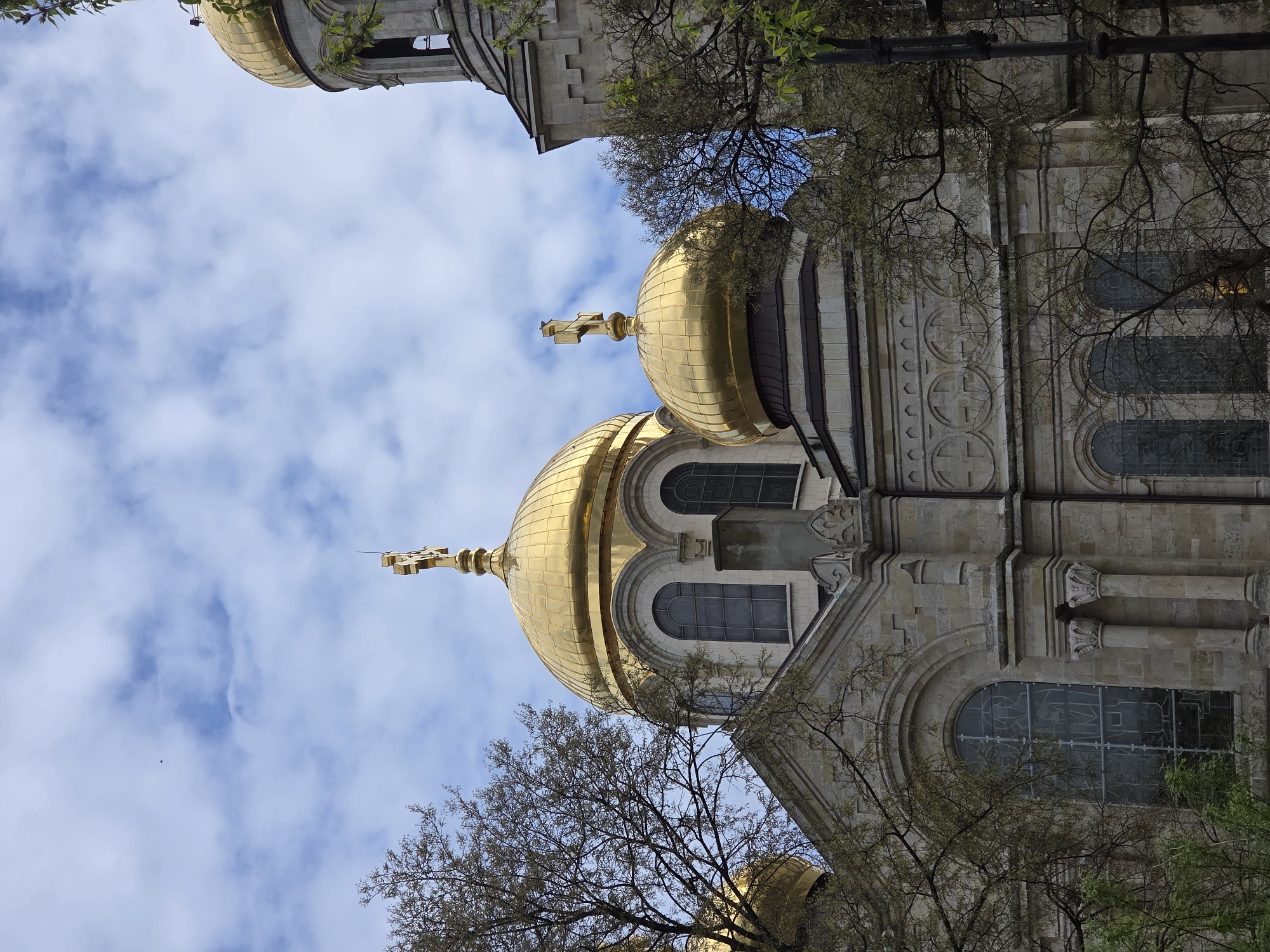 The image shows the upper portion of the Dormition of the Theotokos Cathedral in Varna, Bulgaria. It features the distinctive golden domes topped with Orthodox crosses against a backdrop of a partly cloudy, blue sky. The architectural style is visible in the stone facade, with arched windows and decorative elements. Branches of a tree, with sparse foliage, are visible in the foreground, partially obscuring the view of the cathedral. The photograph is taken from a low angle, emphasizing the height of the structure.