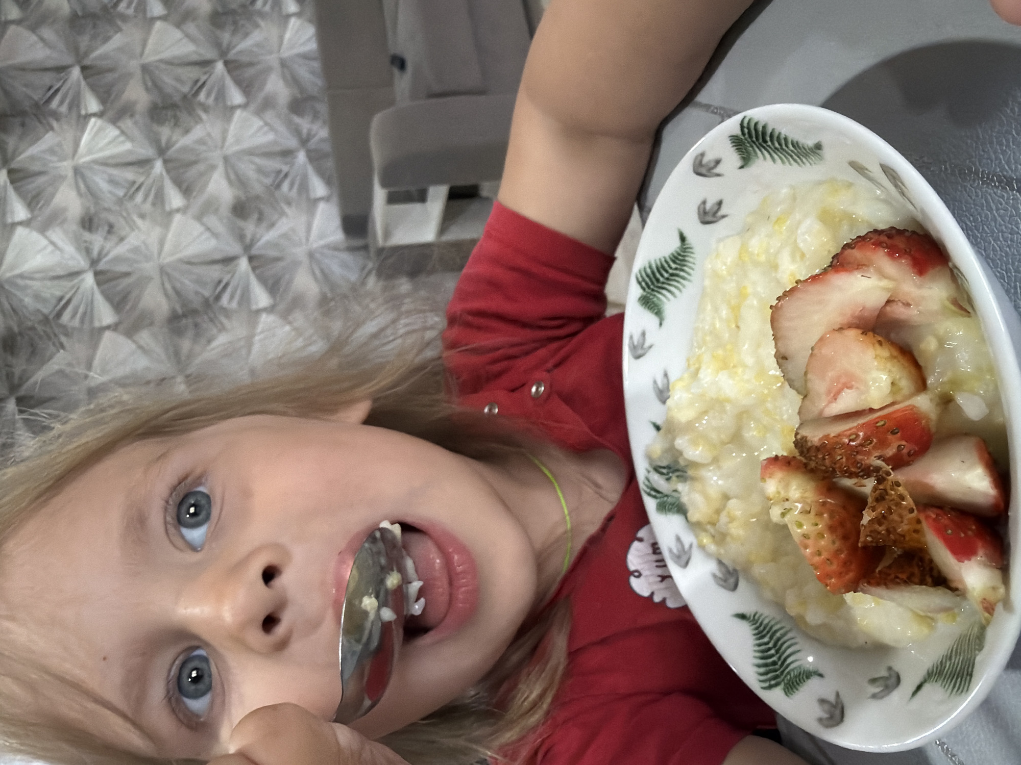 A young girl with blonde hair is eating porridge with strawberries from a bowl. She is wearing a red shirt and is being fed by a spoon held by an unseen person. The background shows a textured wallpaper and a sofa in the distance. The image appears to be taken indoors, possibly in a home environment. It is difficult to determine the exact location in Chaikovsky, Russia, from the provided image alone.