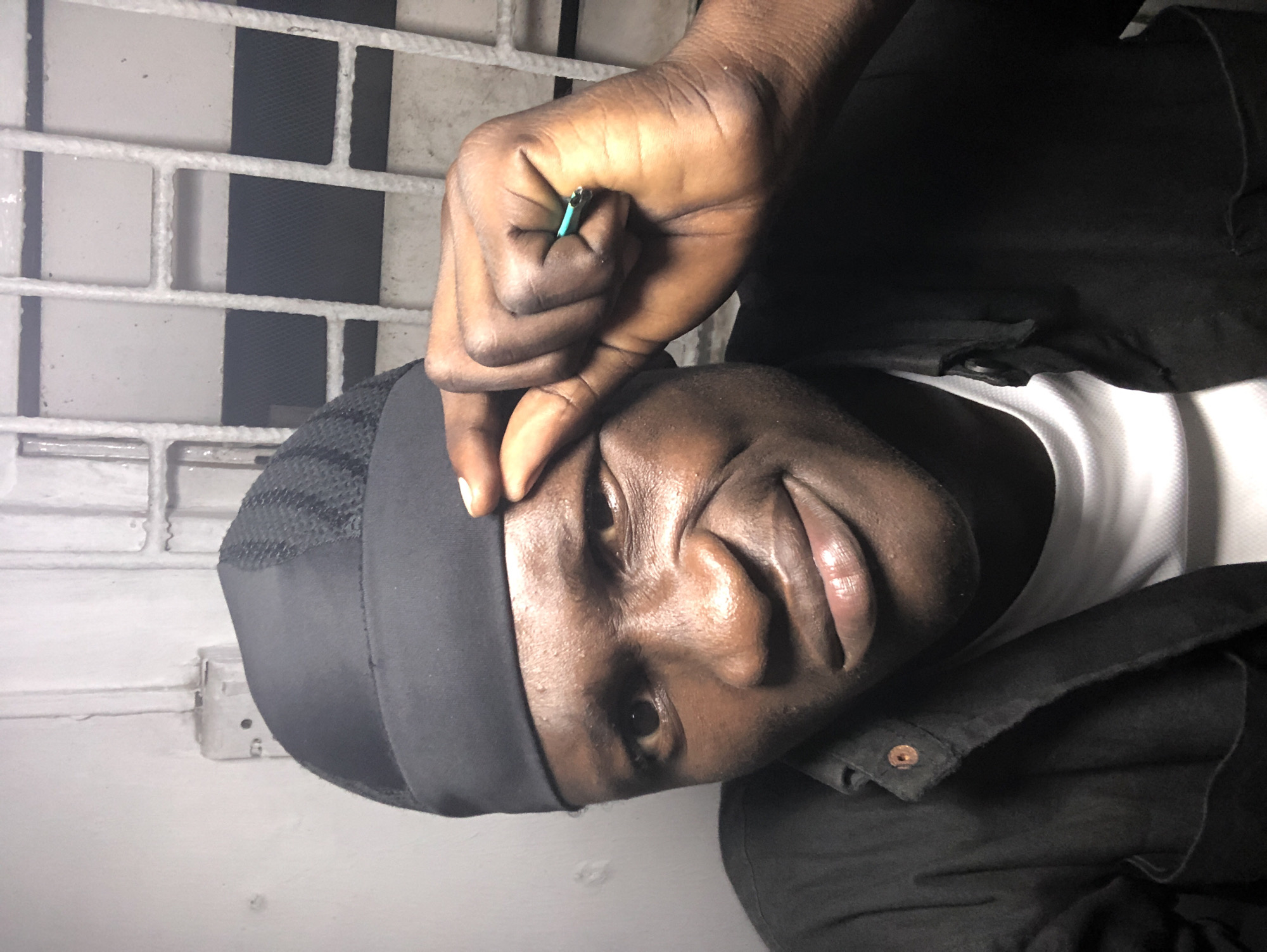 A medium shot captures a man from the chest up, positioned in what appears to be a dimly lit room with a gridded window behind him, suggesting an urban setting. The man is looking towards the camera with a slightly squinted eye and a subtle smile. He is wearing a black durag and a black button-up shirt over a white t-shirt. His right hand is raised, with his index finger and thumb pinching the brim of his durag, and a small, possibly turquoise, object, likely a pen cap, is visible between his fingers. The background shows a light-colored wall with some textural imperfections and a barred window, which is a common sight in many urban environments in Nigeria, including Ado. The lighting is artificial, casting shadows and highlighting the contours of his face. The overall impression is a casual, perhaps candid, photograph taken indoors.