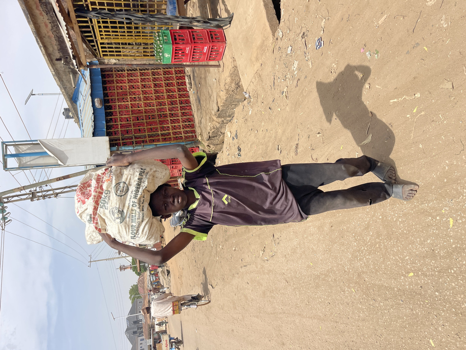 The image, depicting a vertical scene but rotated on its side, captures a young man carrying a heavy sack on his head on a dusty street in Chikaji, Nigeria. Positioned centrally, the man looks towards the camera with a composed expression and a slight smile. He is dressed in a dark short-sleeved t-shirt with green accents and the visible text "SPOR," paired with dark, worn trousers and simple sandals. His hands are raised to support the large, white sack, which bears prominent text including "25KG," "FLOUR MILLS OF NIGERIA PLC," and the address "Old Dock Road, Apapa, Lagos, Nigeria," indicating its origin and likely contents as flour or grain.
The setting appears to be a bustling street or market area under bright, clear daylight, evident from the strong shadows cast by the man and other elements on the sandy ground. In the background, structures resembling shops or stalls feature metal grates and a blue awning. Stacks of red plastic crates, clearly branded "Coca-Cola," are visible near one building. Power lines and poles extend across the sky on the left. Further down the street, faint figures of other people, including one on a bicycle, suggest routine daily activity. The scene conveys a sense of everyday life and manual labor in a vibrant, sunlit environment.