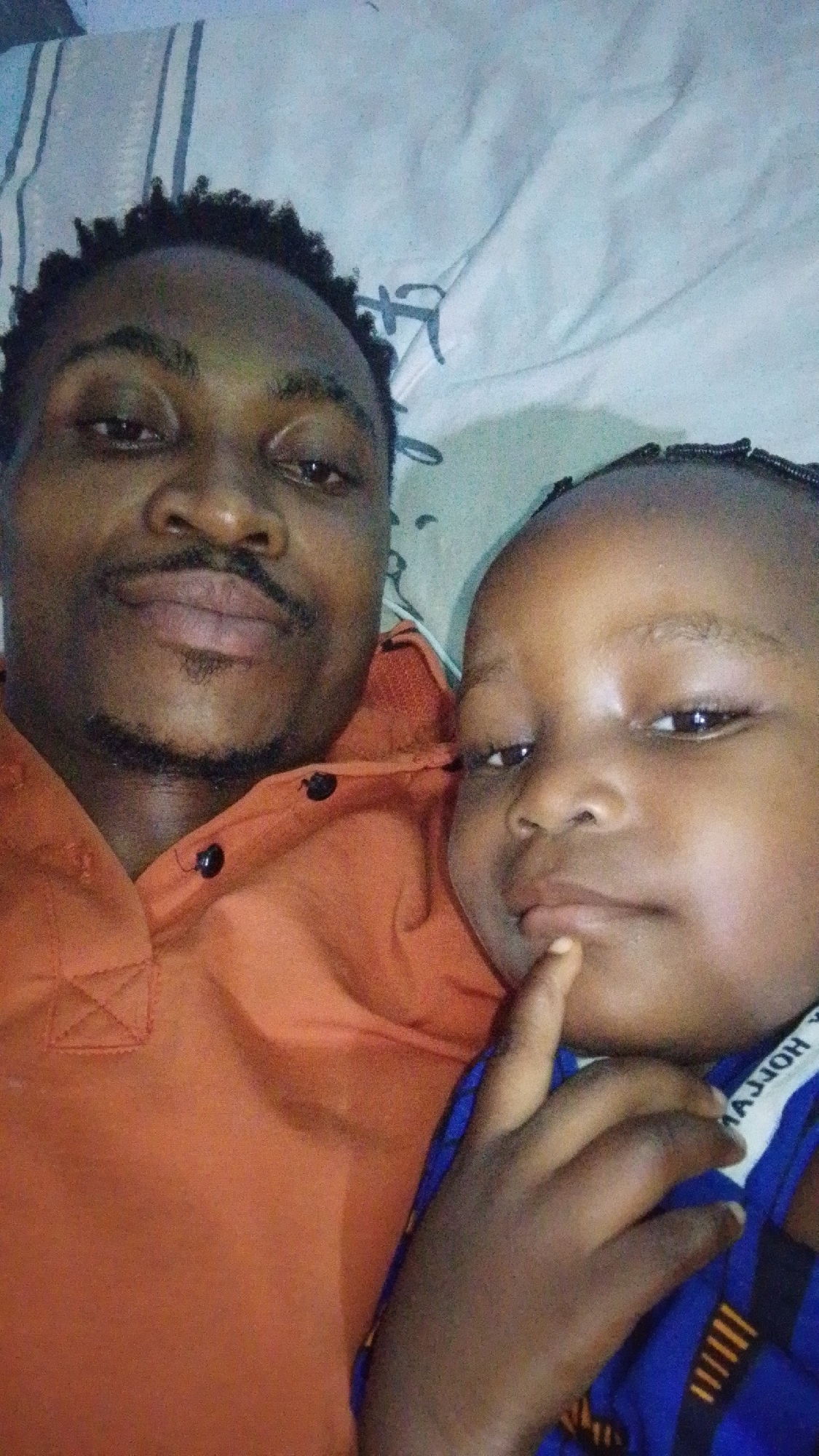 A close-up selfie shows a man and a young child lying down, likely on a bed. The man, positioned on the left, is looking towards the camera with a faint smile. He has short, dark, curly hair and a goatee. He is wearing an orange collared shirt with dark buttons. The child, on the right, is looking slightly away from the camera and has a finger to their lips. The child is wearing a blue shirt with white trim that has some lettering. The background appears to be a light-colored surface with some patterned designs, possibly a duvet or blanket. The scene is indoors and the lighting suggests it was taken with a phone camera. There is no visual information in the image to determine the specific location in Adankolo, Nigeria.