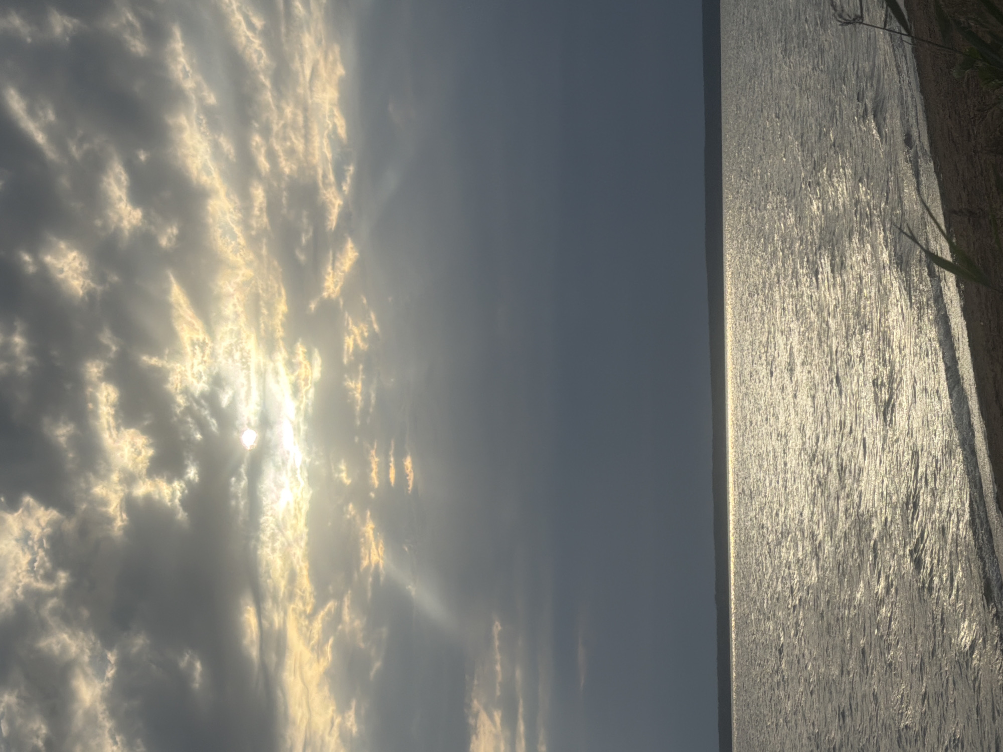 The image depicts a body of water, likely a lake or a wide river, under a dramatic, cloudy sky. The sun is partially obscured by dark clouds, allowing shafts of light to pierce through, creating a dappled effect on the water's surface. The water is rippled, reflecting the sunlight. A dark, thin line of land is visible on the horizon, separating the water from the sky. The foreground shows a sandy shore with some sparse vegetation. The overall atmosphere is somber and possibly indicative of an approaching storm. The location is specified as Vyazy, Russia.