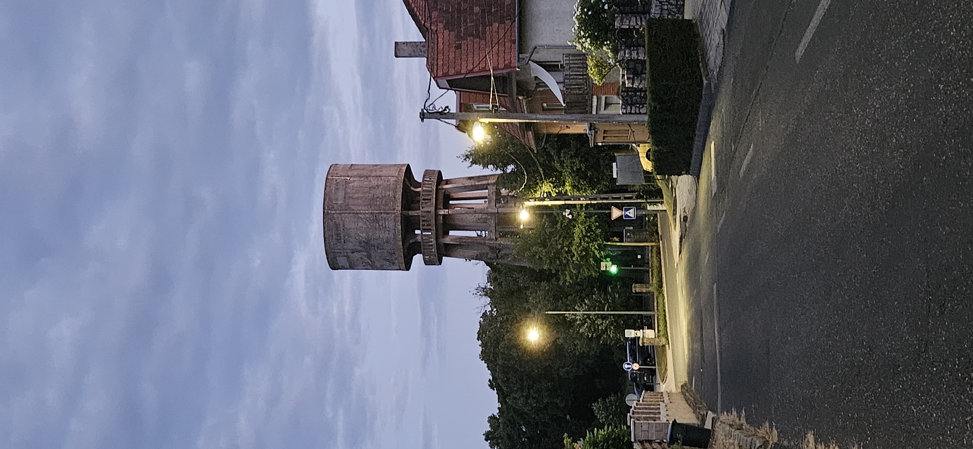 The image depicts a street scene in Hévíz, Hungary, with a prominent concrete water tower as the central subject. The water tower, a tall cylindrical structure supported by pillars, stands against a cloudy twilight sky. The surrounding environment includes residential buildings with red tiled roofs, lush green trees, and streetlights illuminating the asphalt road. Traffic signs and a pedestrian crossing are visible along the street, indicating a residential or semi-urban area. The overall atmosphere is serene and quiet, characteristic of a dusk or early evening setting in a European town.