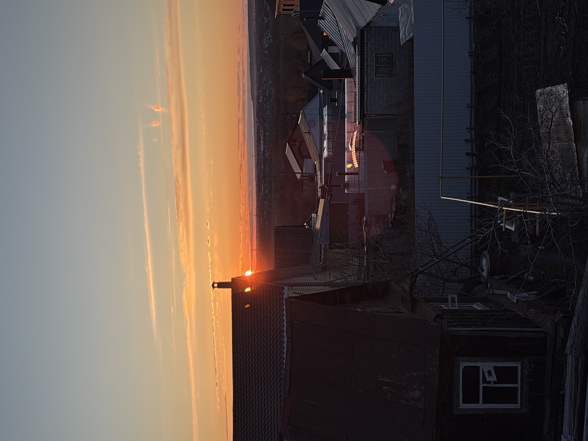 The image, when viewed correctly (rotated 90 degrees clockwise), captures a tranquil sunset over what appears to be a semi-urban or rural landscape, set in Ulyanovsk, Russia. The time of day is dusk, with the sun low on the horizon, casting an intense orange and yellow glow across the sky and reflecting on cloud formations. The sky transitions from vibrant warm colors near the horizon to a softer, pale blue overhead.
In the foreground and midground, several buildings with various roof styles (including corrugated metal and shingle-like materials) are silhouetted against the bright sky. The setting sun is partially obscured by a chimney on one of the roofs, creating a bright highlight. Bare trees and bushes, suggesting late autumn or winter, are visible, particularly on the right side of the frame, intertwining around the structures. One building features a distinctive white-framed window with a pane slightly ajar. Pipes or utility lines run across some areas.
The background shows a distant, hazy landscape, likely a town or city, stretching across the horizon. Two small, bright, somewhat rectangular flares or reflections are visible in the upper sky, possibly atmospheric phenomena or lens effects. The scene evokes a sense of quietude and the peaceful transition from day to night. There are no people visible, and no specific activity is taking place. No discernible text is present within the image.