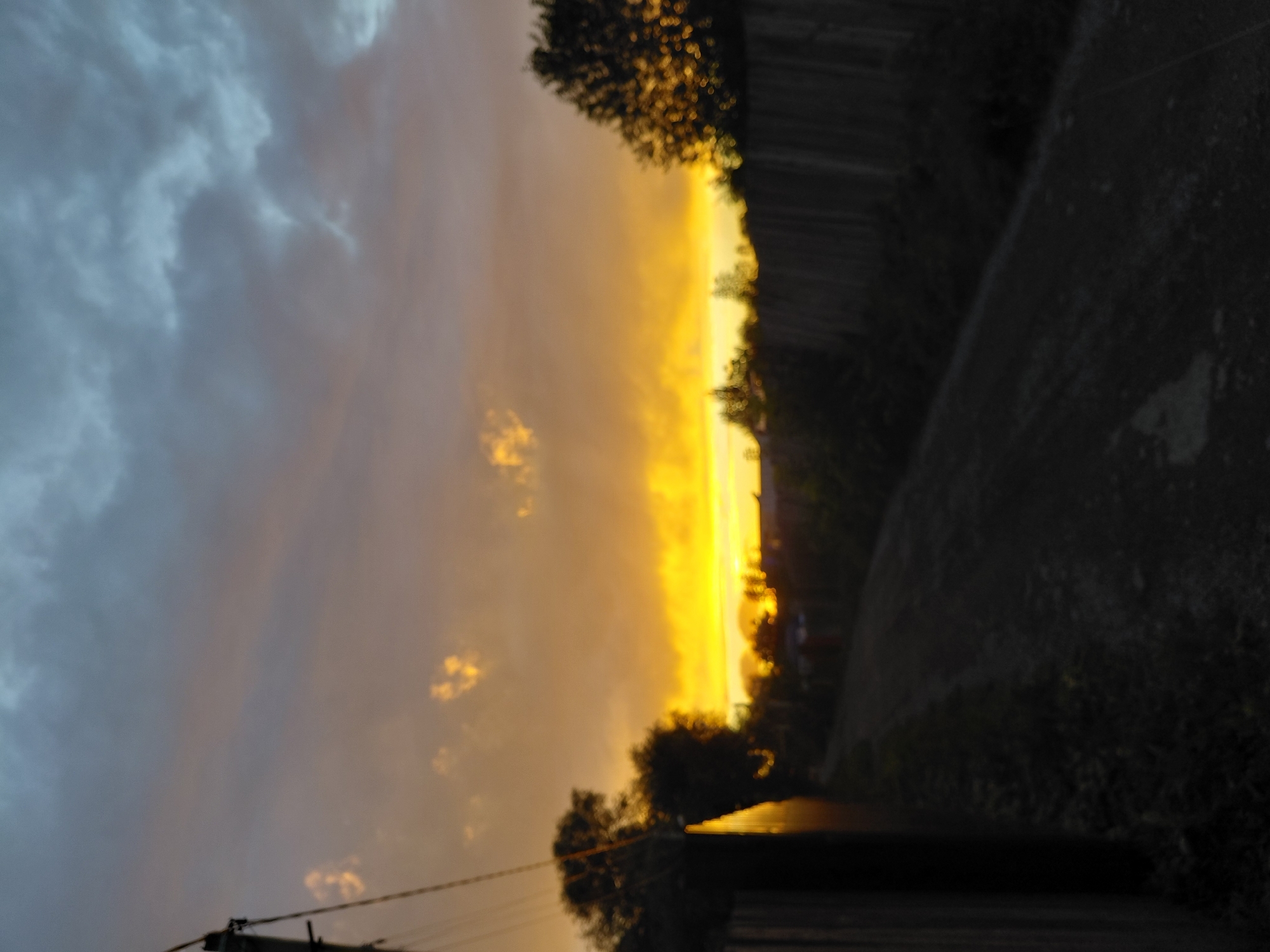 This is a photograph of a dirt road in a rural or suburban area at dusk. The sky is filled with dramatic clouds, with the lower portion illuminated by the setting sun in vibrant shades of orange and yellow. The upper part of the sky is a darker, muted blue-gray.
On the left side of the image, a wooden fence runs along the road, partially obscuring a pole with wires. Silhouetted trees are visible behind the fence, with some of their foliage catching the warm light of the sunset, creating a bokeh effect. On the right side of the road, another wooden fence is visible, also running parallel to the road. More trees and vegetation are present, some of which are also silhouetted against the bright sky. The road itself appears to be unpaved, with a rough texture. The overall atmosphere is serene and suggests the end of the day in a quiet location.
