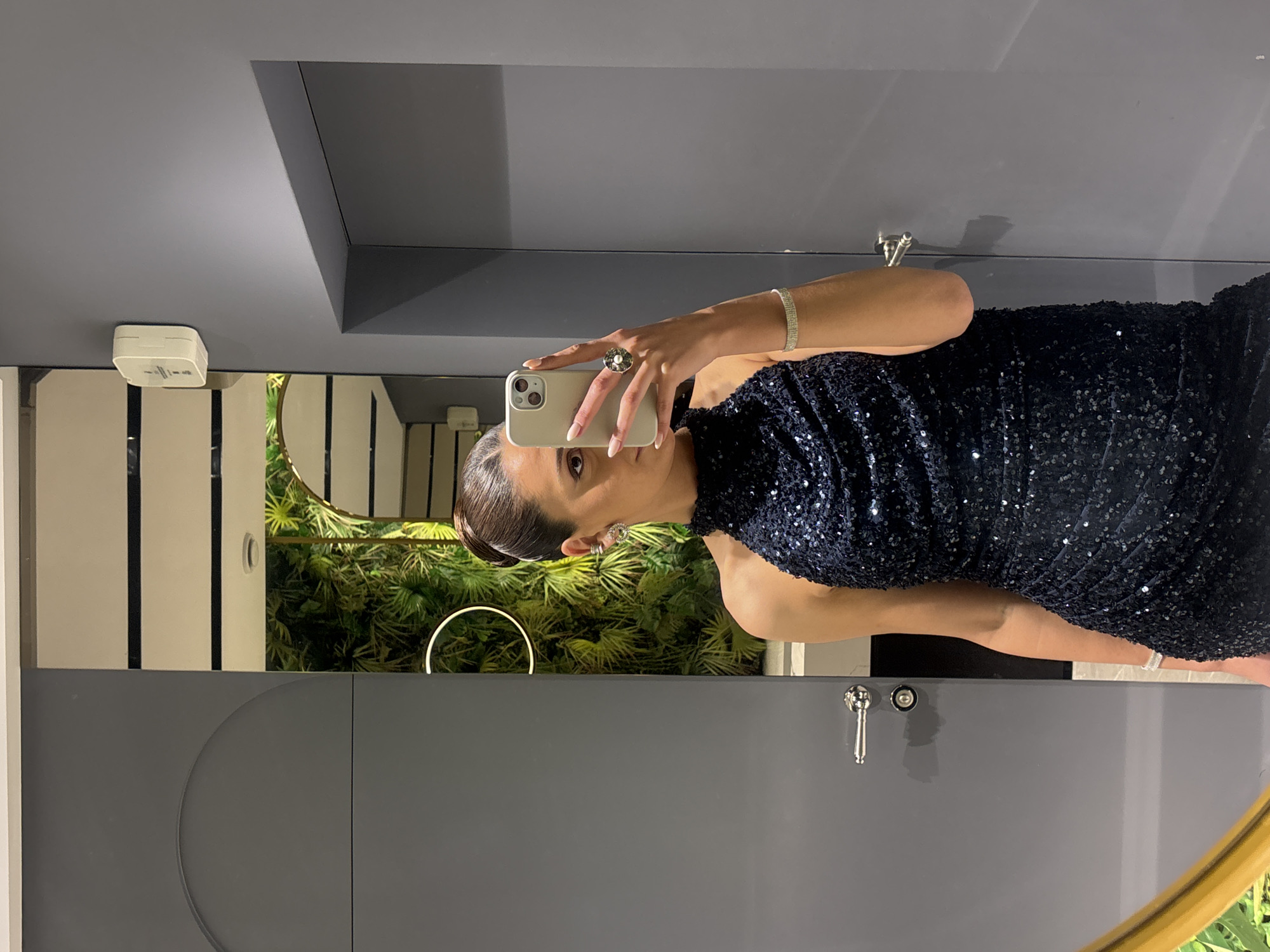 A woman is standing in a modern bathroom, taking a selfie in the mirror with her iPhone. She is wearing a sleeveless, dark blue dress with sequins, a ring on her left hand, and a bracelet on her right wrist. The bathroom has gray walls, and in the mirror's reflection, there is a green plant. The location is most likely a private residence in the city of Masis, Armenia.