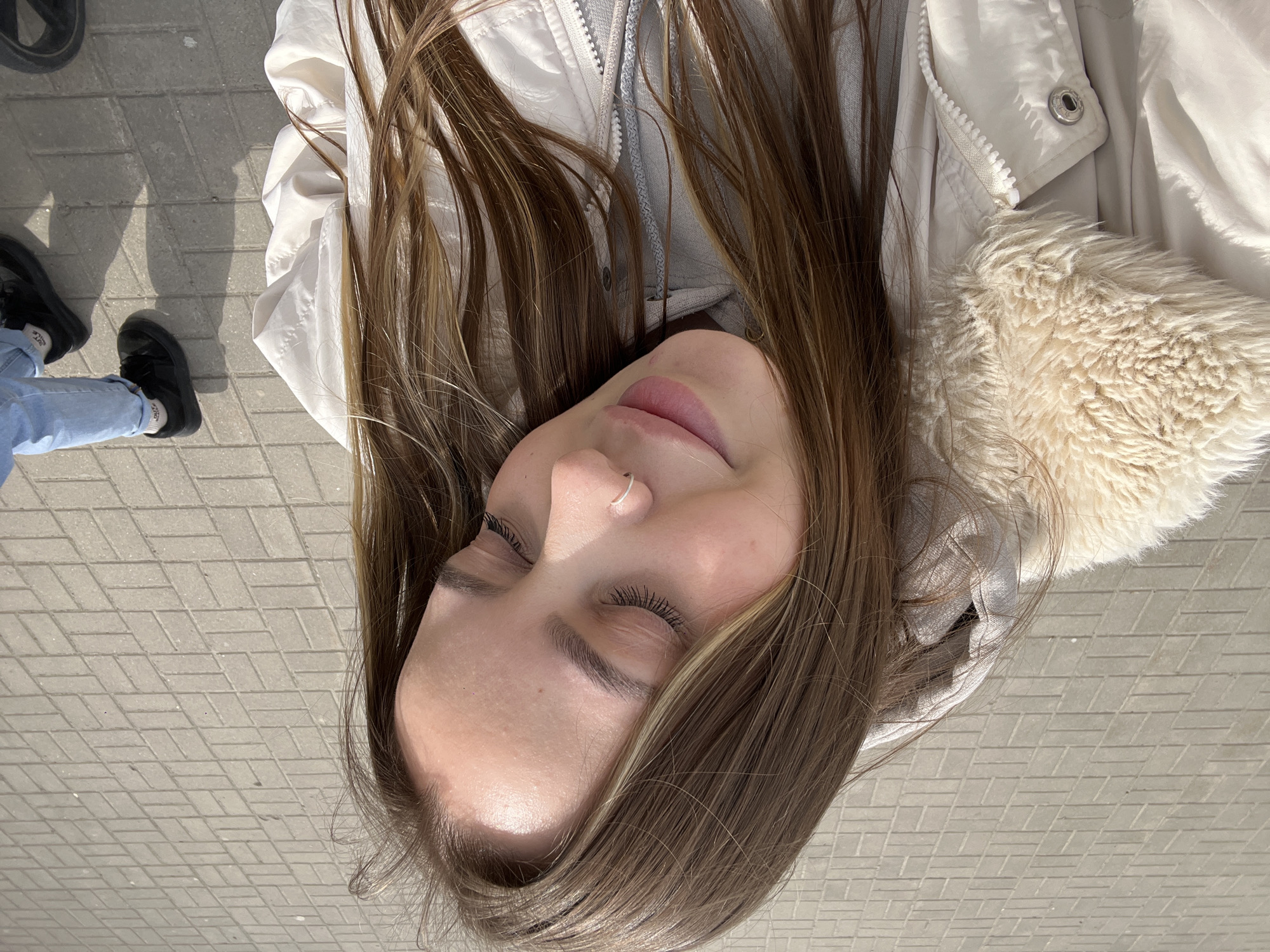 A woman is lying on a tiled surface, possibly a sidewalk or paved area. She is wearing a light-colored jacket, and her hair is spread around her head. Part of a person wearing jeans and black shoes is visible in the background. The location is not determinable from this image, and a specific location in Novopokrovskaya, Russia cannot be identified.