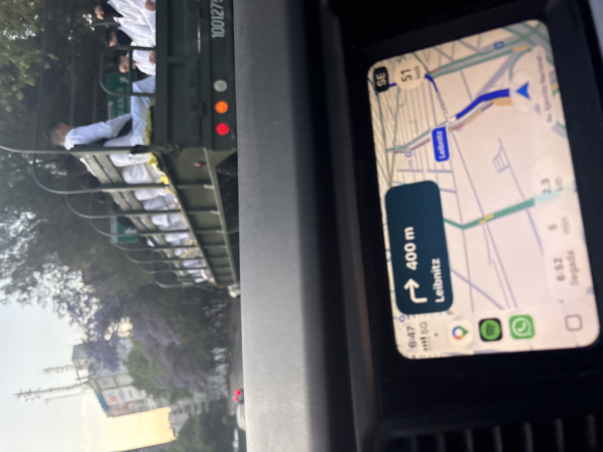 The image, rotated 90 degrees, captures a street scene from inside a car in Mexico City. The viewer is driving behind a military-style truck while referencing a navigation screen.
On the right side of the frame, a smartphone or car display shows a map application. The map indicates a direction to turn left in "400 m" onto "Leibnitz" street. A compass icon points "SE," and a speed of "51" (likely km/h) is displayed. The current time shown is "6:47," with an estimated arrival time ("llegada") of "6:52." Below the map, icons for Google Maps, Spotify, and WhatsApp are visible.
To the left, an olive green, military-style truck is visible, with multiple individuals standing in its open bed. Many of these individuals are wearing white uniforms, possibly indicating medical personnel, students, or service members. One person in a white uniform is leaning over the side of the truck. The truck's rear features red and amber lights, and a partial numerical marking "1001275" is visible on its side.
In the background, lush trees with distinctive purple flowers, consistent with jacaranda trees common in Mexico City, line the street. Buildings are partially visible through the foliage. The time shown on the navigation screen suggests either early morning or late afternoon, and the weather appears clear. The scene depicts an urban drive, with the viewer following the truck through the city streets.