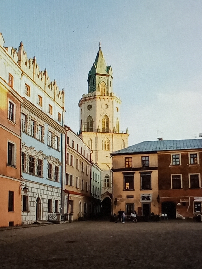 The image displays a daytime view of the Rynek (Market Square) in Legnica, Poland. Dominating the background-center is the Old Town Hall tower (Stary Ratusz), a multi-tiered, light-colored structure with gothic-style arched windows, topped by a pointed green spire.
To the left, a row of multi-story historic tenement buildings lines a street or passage. The building closest on the left features an orange-brown base, a light blue mid-section with ornate window surrounds, and a white upper level with a decorative stepped gable. A black wall-mounted lantern is visible on its lower facade. Further down this row, buildings with simpler beige and light brown facades recede.
On the right side of the frame, two multi-story buildings face the square. The left building is light brown/ochre with dark windows and a sign reading "ONE" above an entrance. The adjacent building to its right is darker brown.
The foreground consists of a dark, paved square, likely cobblestone, with visible textural variations and patches of shadow extending from the right, suggesting directional sunlight. A small group of approximately three to five indistinct figures is present near the base of the buildings on the right, possibly interacting or seated. The sky is clear and light blue.