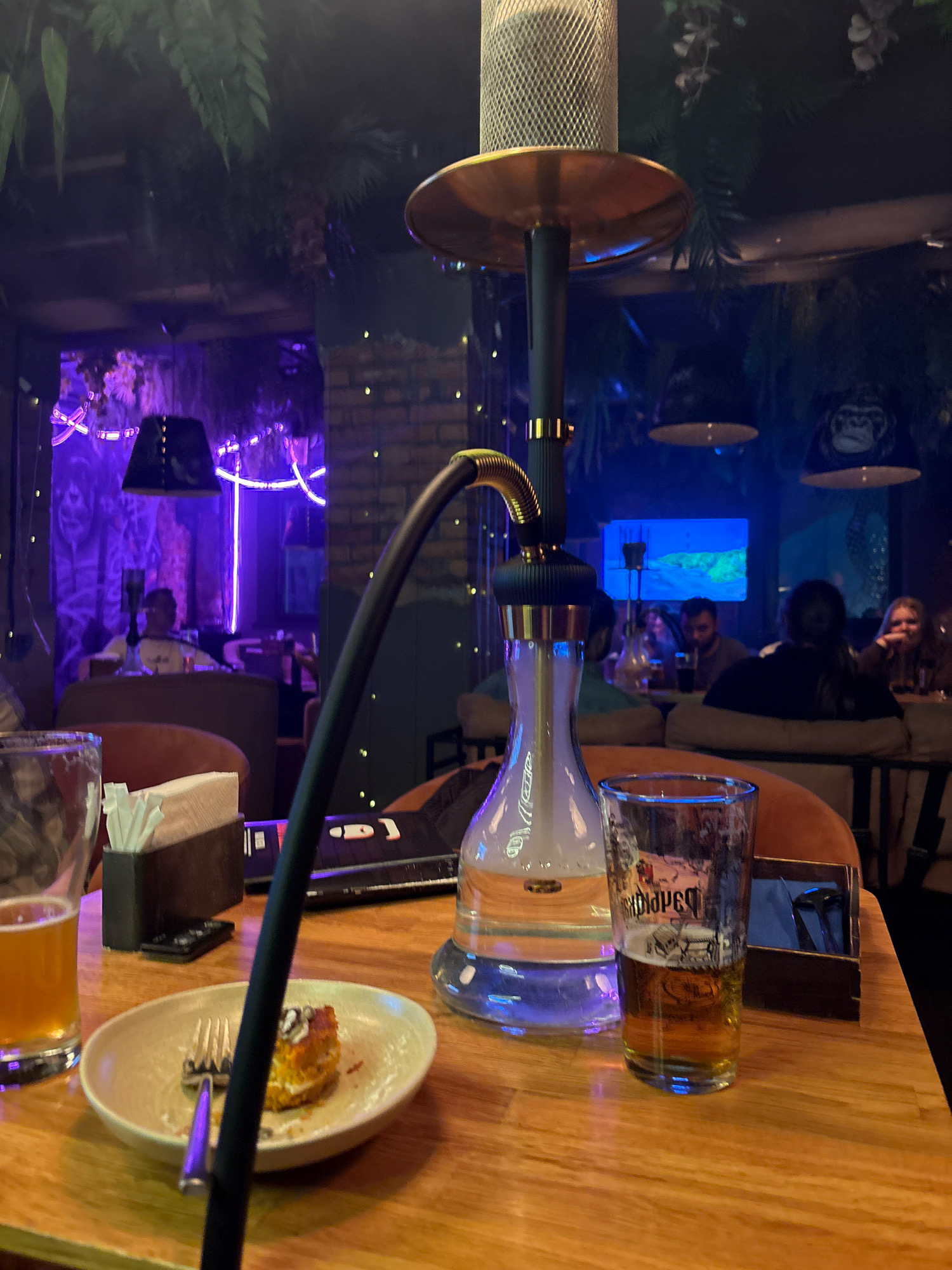 The image depicts an indoor hookah lounge setting with a dimly lit, atmospheric ambiance. In the foreground, a wooden table holds a partially consumed dessert on a plate, a fork, a glass of amber-colored liquid, and a partially filled glass with a logo visible. A hookah with a glass base filled with liquid and smoke, a black stem, and a metal bowl is centrally positioned. A black hose extends from the hookah towards the left. The background reveals a lounge area with people seated at tables, dimly illuminated by purple and blue lights. Decorative elements like plants and images of monkeys are visible on the walls and ceiling. The establishment appears to be a bar or lounge, likely in Minsk, Belarus, given the context provided.