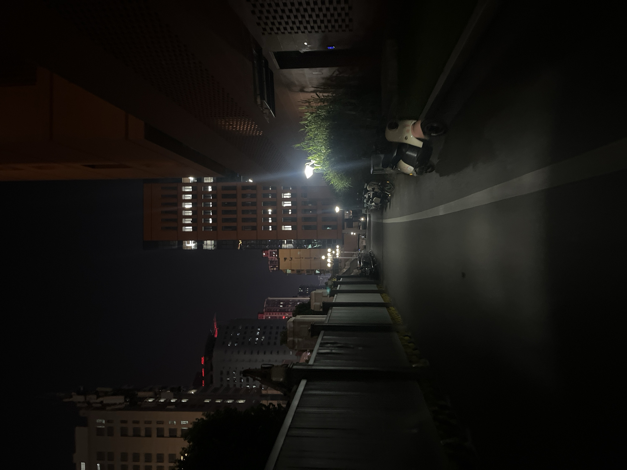 The image captures an urban nocturnal scene in Yuzhong District, China, presenting a low-angle view down a paved pathway. On the left, a dark, slatted fence with periodic lighter pillars extends into the background, partially obscuring dark green foliage. Beyond the fence, multiple multi-story buildings rise against a black sky, with numerous illuminated rectangular windows visible across their facades. One building displays a distinct tall, segmented vertical structure, and red lights are discernible on distant building rooftops. On the right, a large, multi-story building with a reddish-brown exterior dominates the frame, featuring many illuminated square windows. Its lower section includes textured wall panels and a dark recessed entrance. A bright streetlight casts illumination upon a dense patch of green vegetation positioned along the building's base. Several scooters or small motorcycles are parked on the paved surface adjacent to the vegetation, with a light-colored scooter prominent in the immediate foreground. The dark paved pathway exhibits a continuous white line running longitudinally from the foreground into the receding distance. Additional streetlights provide ambient illumination further down the pathway. No individuals are visible within the scene.