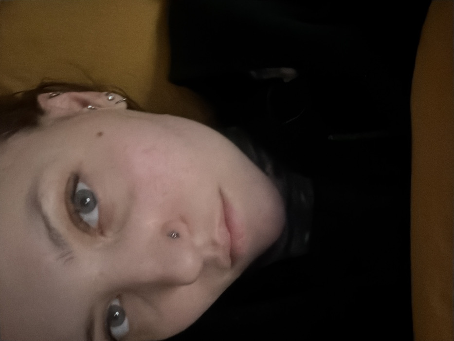 A close-up, tilted portrait reveals a person with fair skin, light eyes, and several ear piercings. They also have a nose piercing. Their facial expression is neutral, and they appear to be lying down, resting against a yellow surface. They are wearing a dark colored shirt or jacket. The background is mostly black.