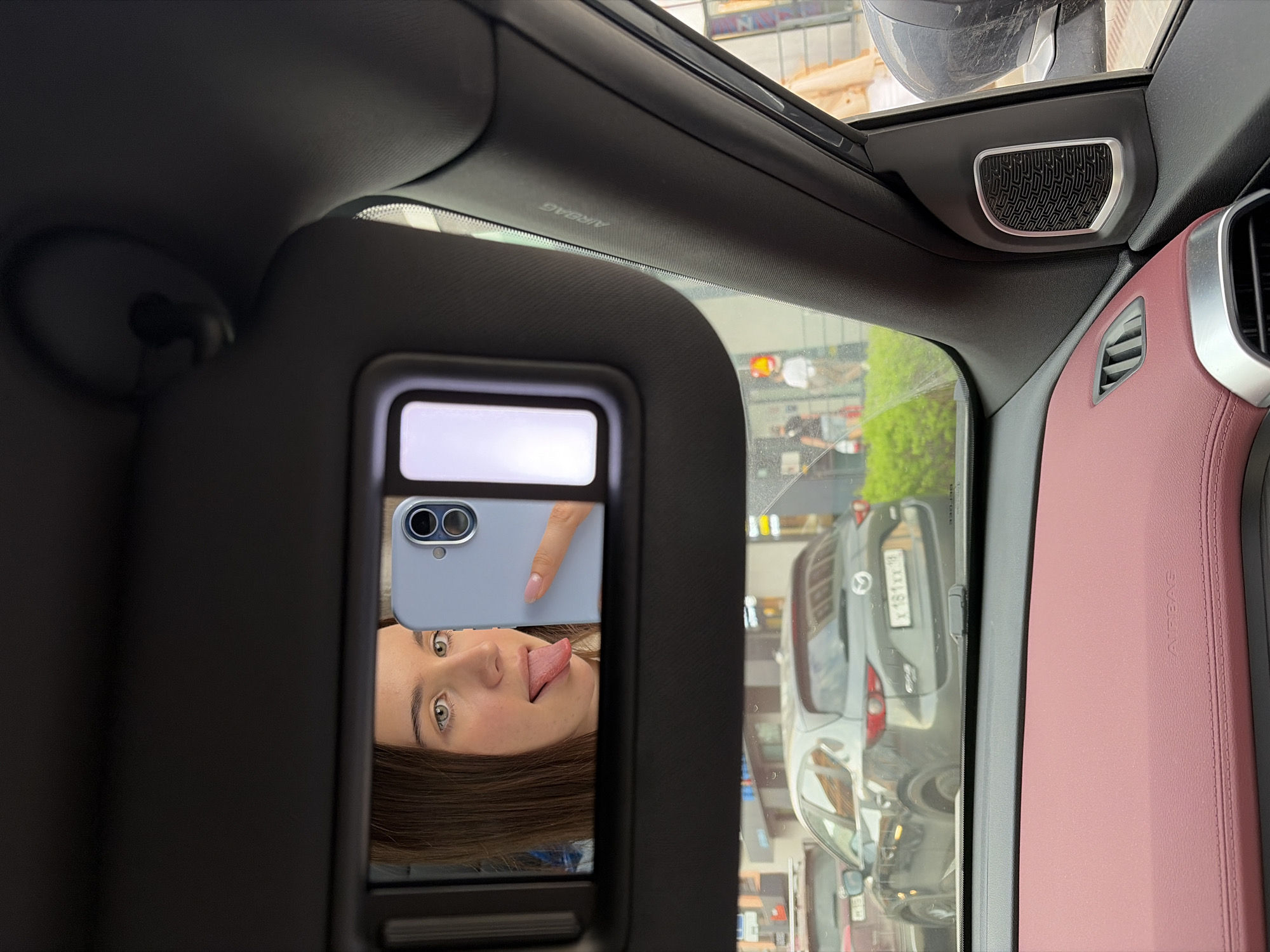 The image, taken from inside a car, shows a rearview mirror reflecting a woman taking a selfie with a smartphone. The woman is making a face, sticking her tongue out. The exterior view seen through the windshield includes a parked vehicle with license plate "X481XX98" likely placing the location in Izhevsk, Russia. The car's interior includes a pink upholstered area with the word "AIRBAG" and a speaker grill.