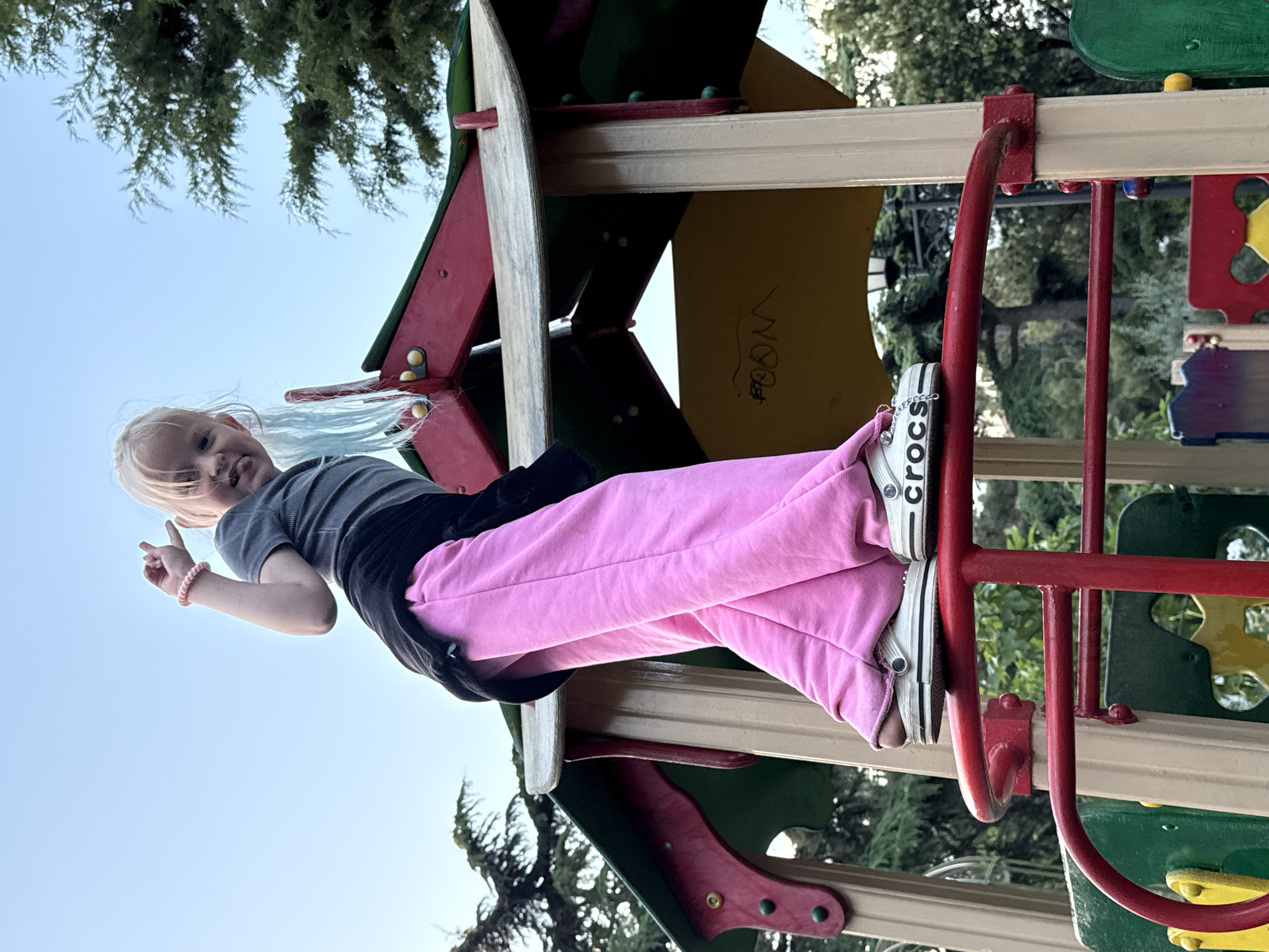 A low-angle shot captures a young girl on playground equipment, seemingly in a park or recreational area. She wears a gray t-shirt, a black wrap or garment around her waist, pink sweatpants, and white Crocs sneakers. She is smiling and holding up two fingers in a peace sign. The playground structure is composed of metal and plastic components in various colors, including red, green, yellow, and beige. Trees surround the playground, visible in the background against a clear blue sky.