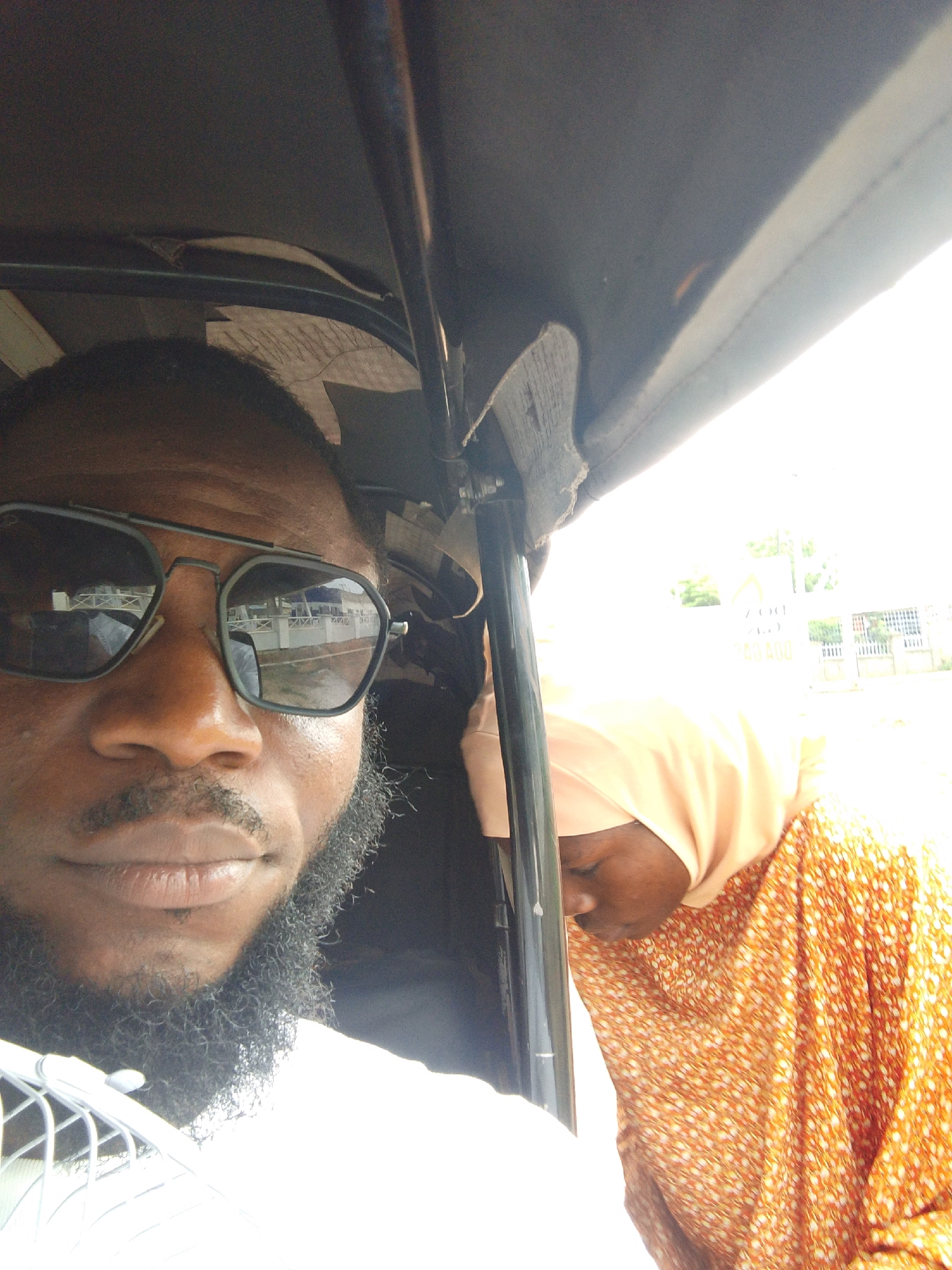 A selfie photograph shows a man with dark skin and a beard, wearing dark sunglasses, inside a vehicle. To the right, a woman wearing a peach-colored hijab and an orange patterned dress is visible. The setting appears to be a three-wheeled vehicle, possibly a "keke" or "auto-rickshaw," commonly used as public transportation. Based on visual cues reflecting in the sunglasses, the location is likely in a built-up area with structures and a bridge, which is consistent with Lokoja, Nigeria. The lighting is bright, suggesting daytime. The vehicle's frame and ceiling are visible, implying the selfie was taken from inside the vehicle.